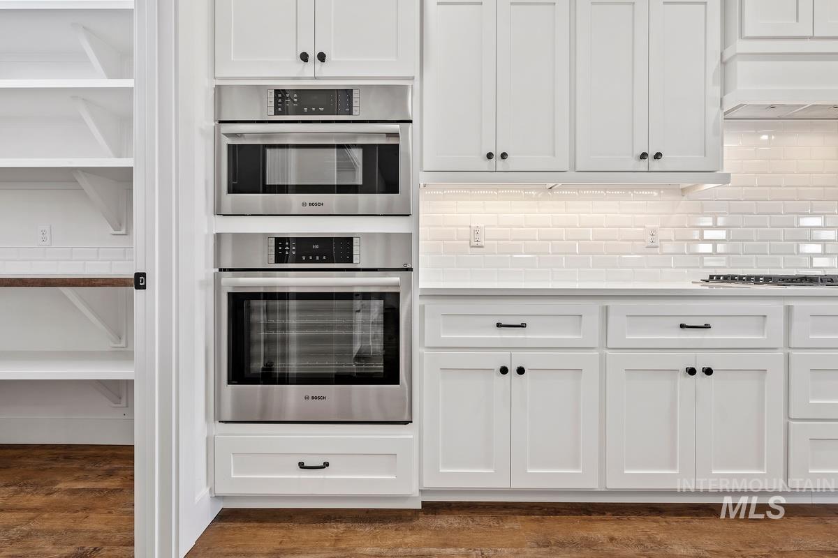 Kitchen featuring white cabinets, dark wood finished floors, double oven, decorative backsplash, and under cabinet range hood