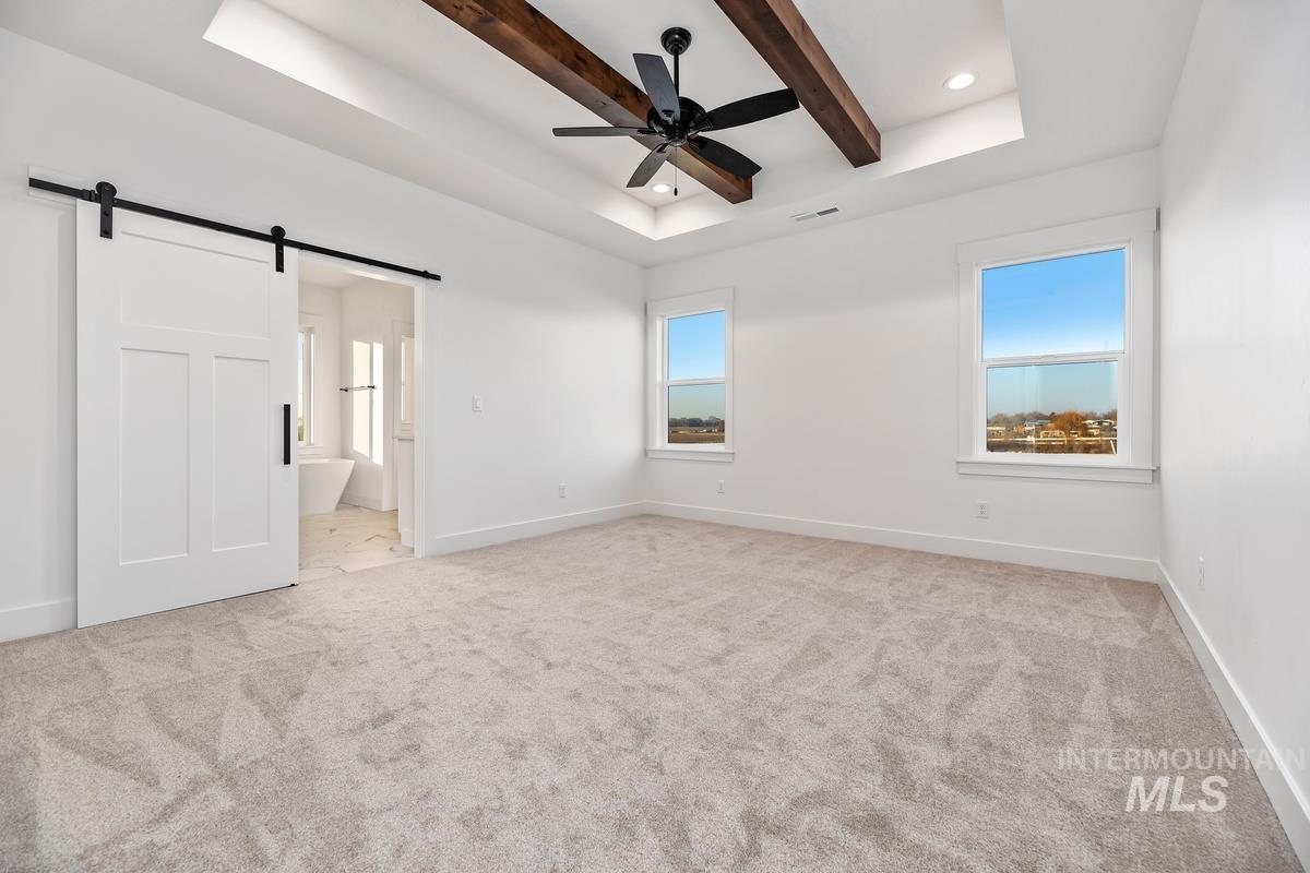 Unfurnished bedroom with a barn door, a raised ceiling, connected bathroom, light carpet, and beamed ceiling