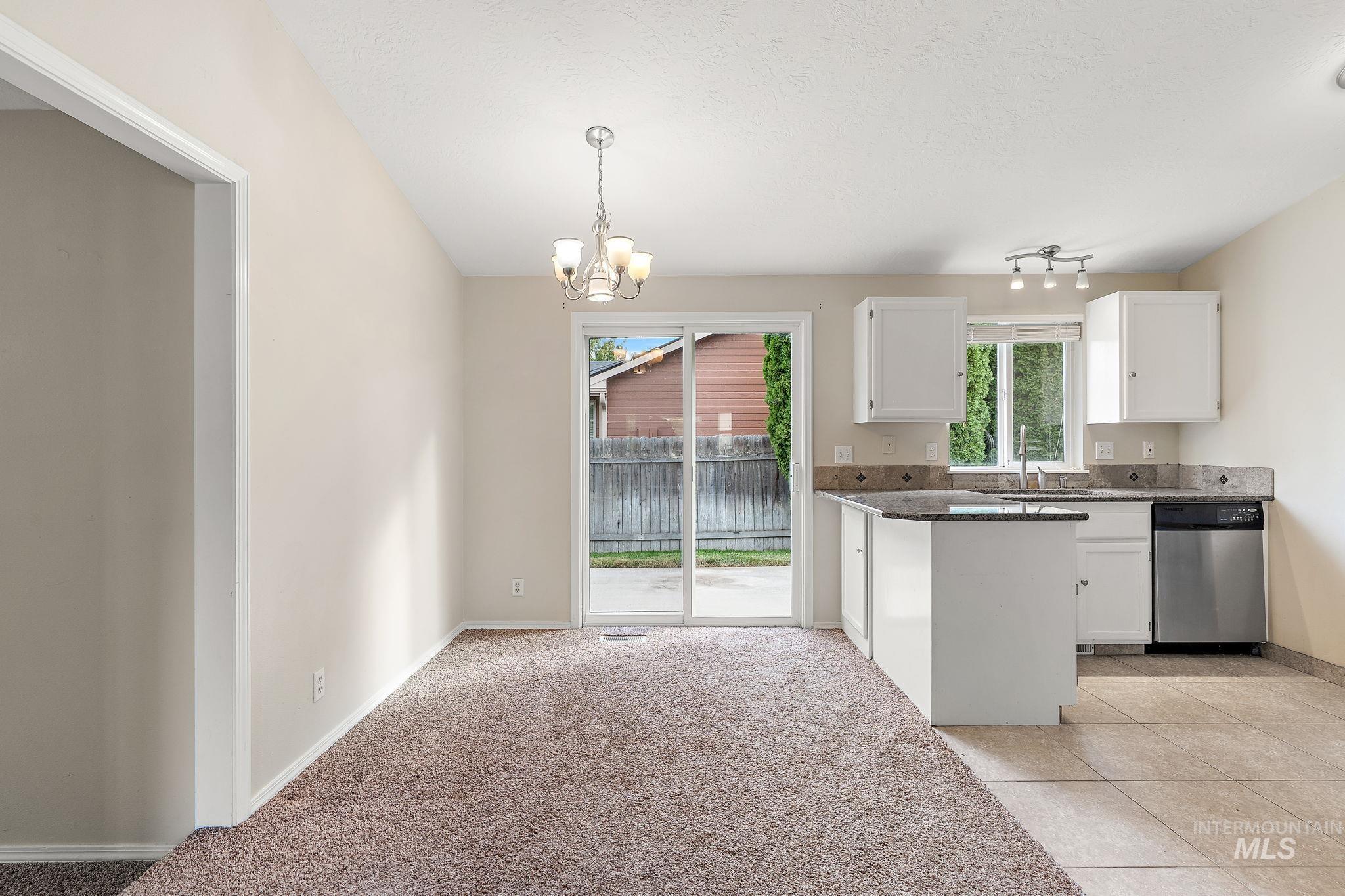 Kitchen featuring white cabinets, stainless steel dishwasher, light tile patterned floors, hanging light fixtures, and dark stone countertops