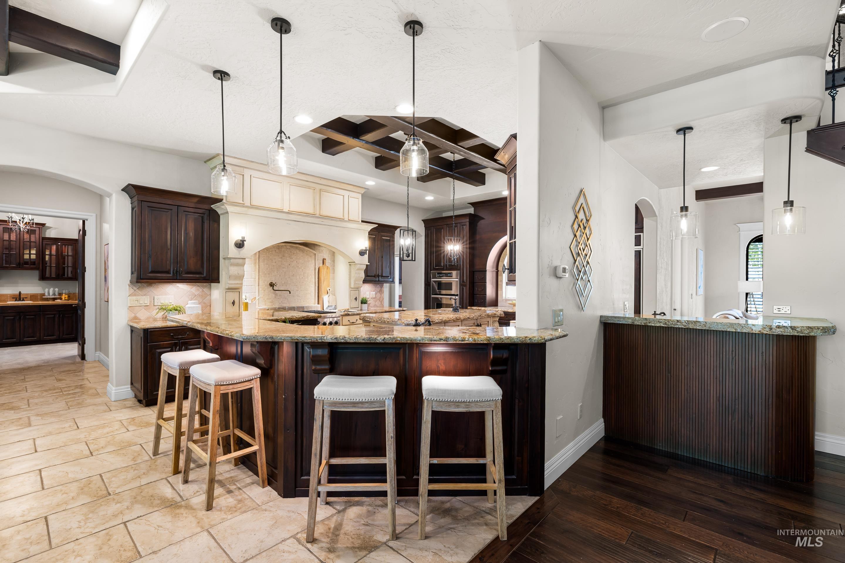 Kitchen with arched walkways, beamed ceiling, coffered ceiling, dark brown cabinets, and light stone countertops