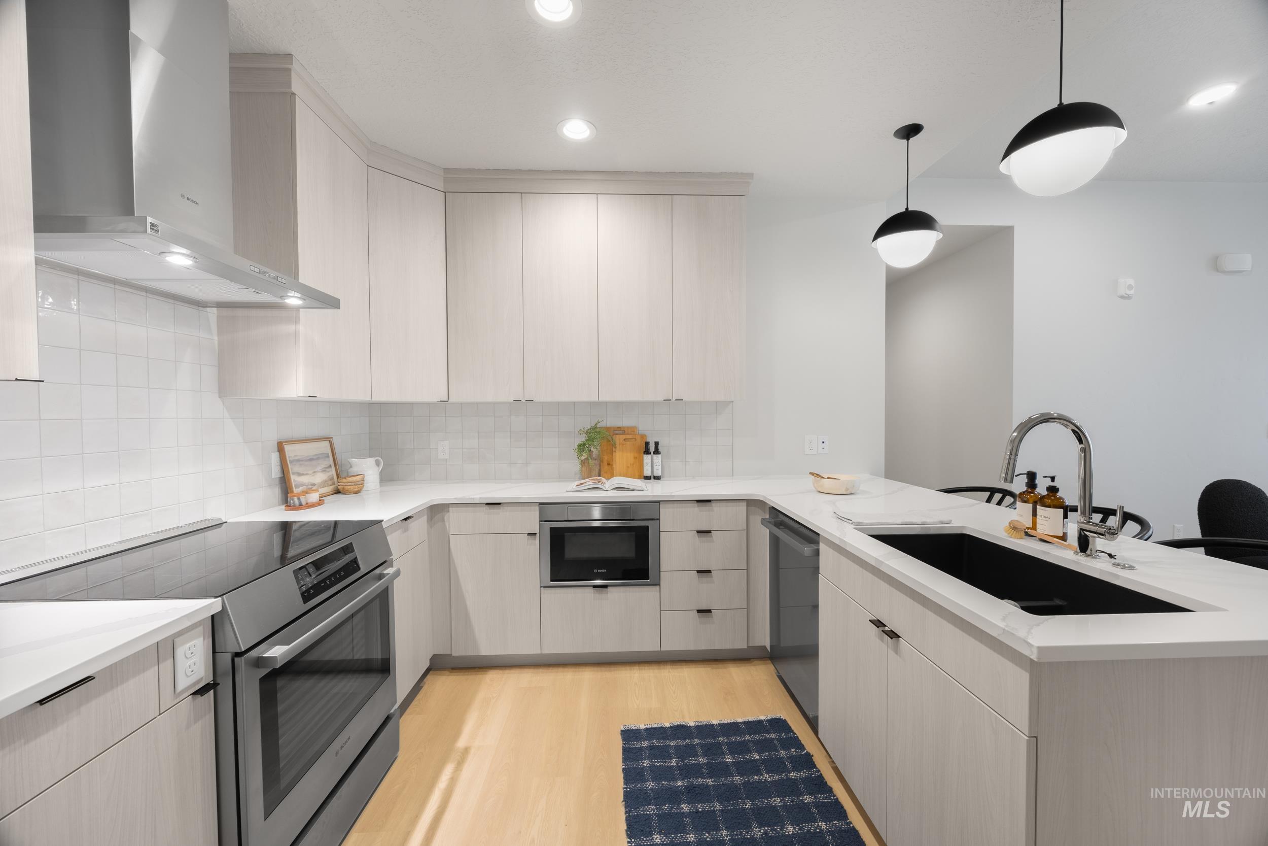 Kitchen with appliances with stainless steel finishes, wall chimney exhaust hood, light wood-style floors, a peninsula, and hanging light fixtures