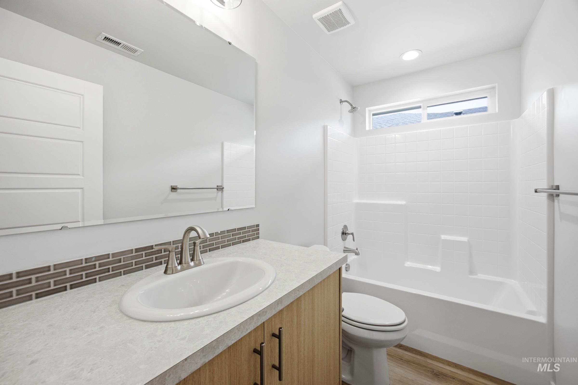 Bathroom featuring a wood-finish vanity with a light-toned countertop, an integrated sink with brushed nickel faucet, a mosaic tile backsplash, and a white tub/shower combination with subway tile-patterned surround