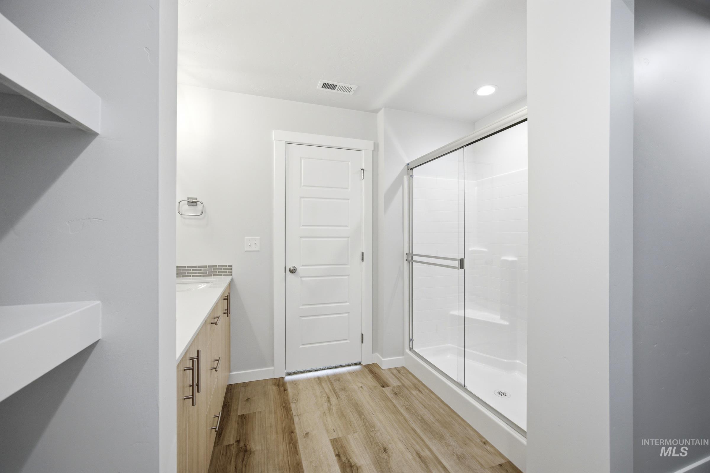 Modern bathroom featuring a wood-finish floor, light wood-tone vanity with a light countertop, mosaic tile backsplash, and a glass-enclosed shower with a built-in bench