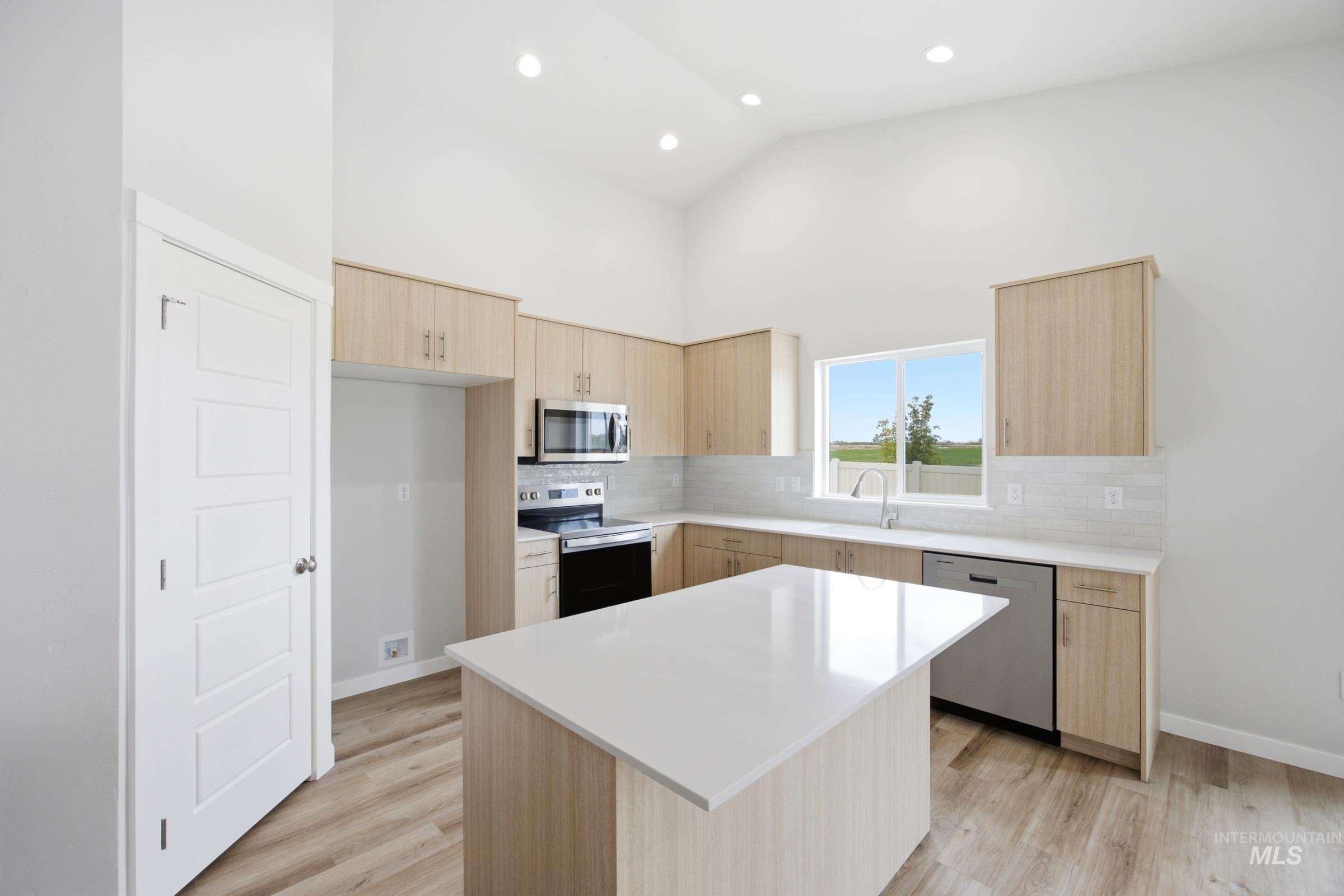 Modern kitchen featuring a central island with white countertop, recessed lighting, wood-finish flooring, light wood cabinetry, and stainless steel appliances