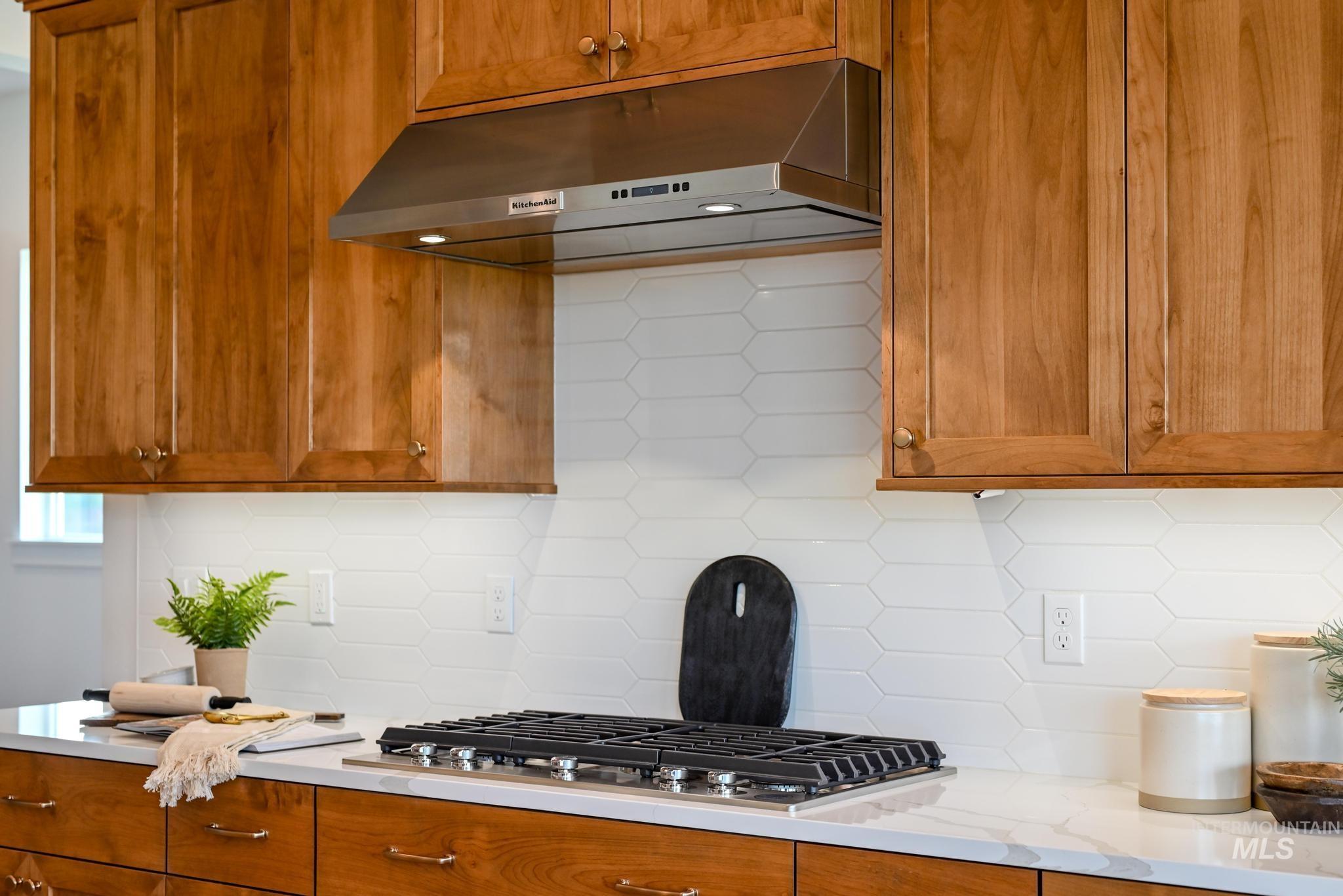 Kitchen with decorative backsplash, brown cabinets, under cabinet range hood, stainless steel gas stovetop, and light stone countertops
