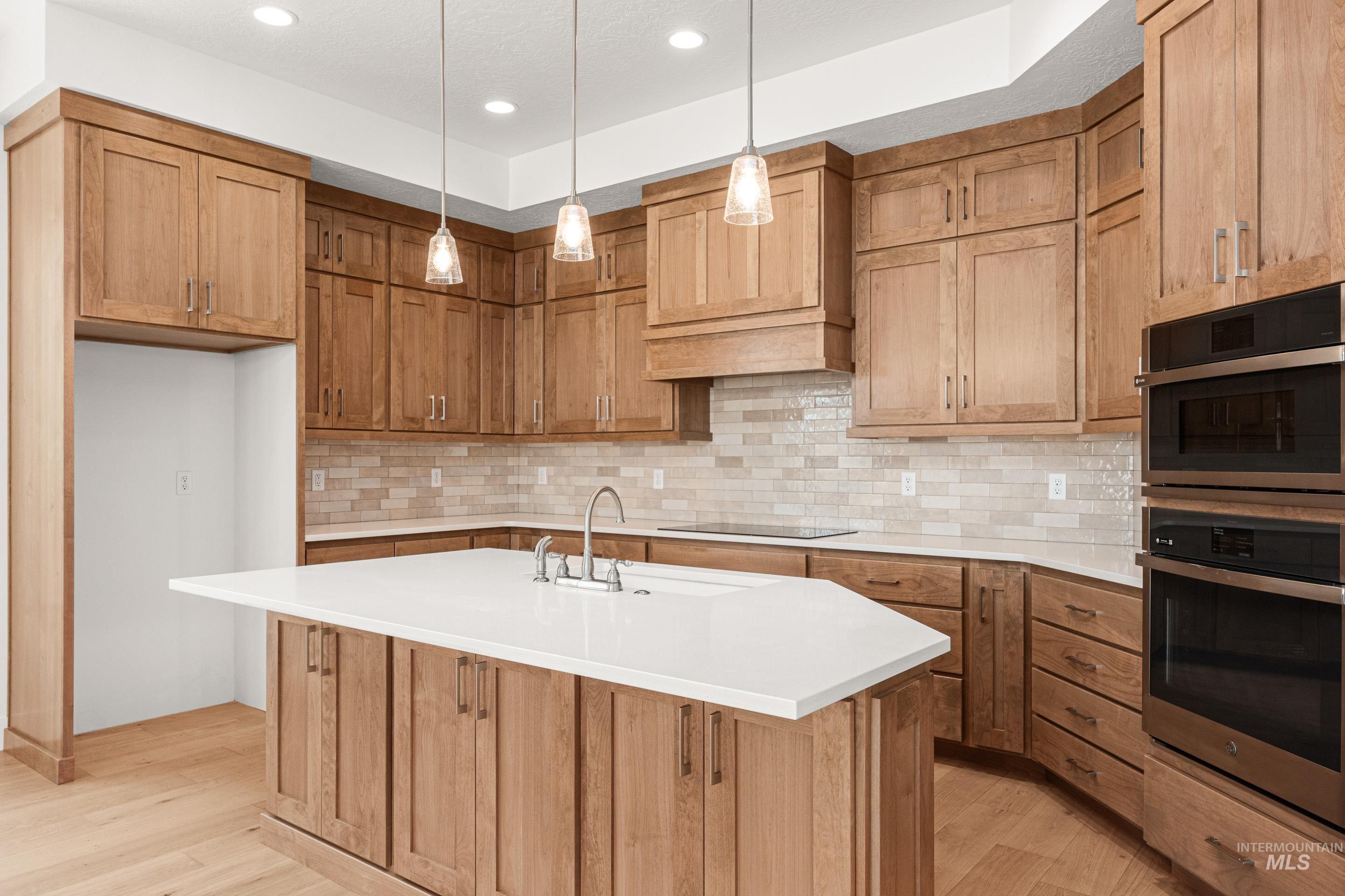 Kitchen with stainless steel double oven, hanging light fixtures, tasteful backsplash, a kitchen island with sink, and light wood-type flooring