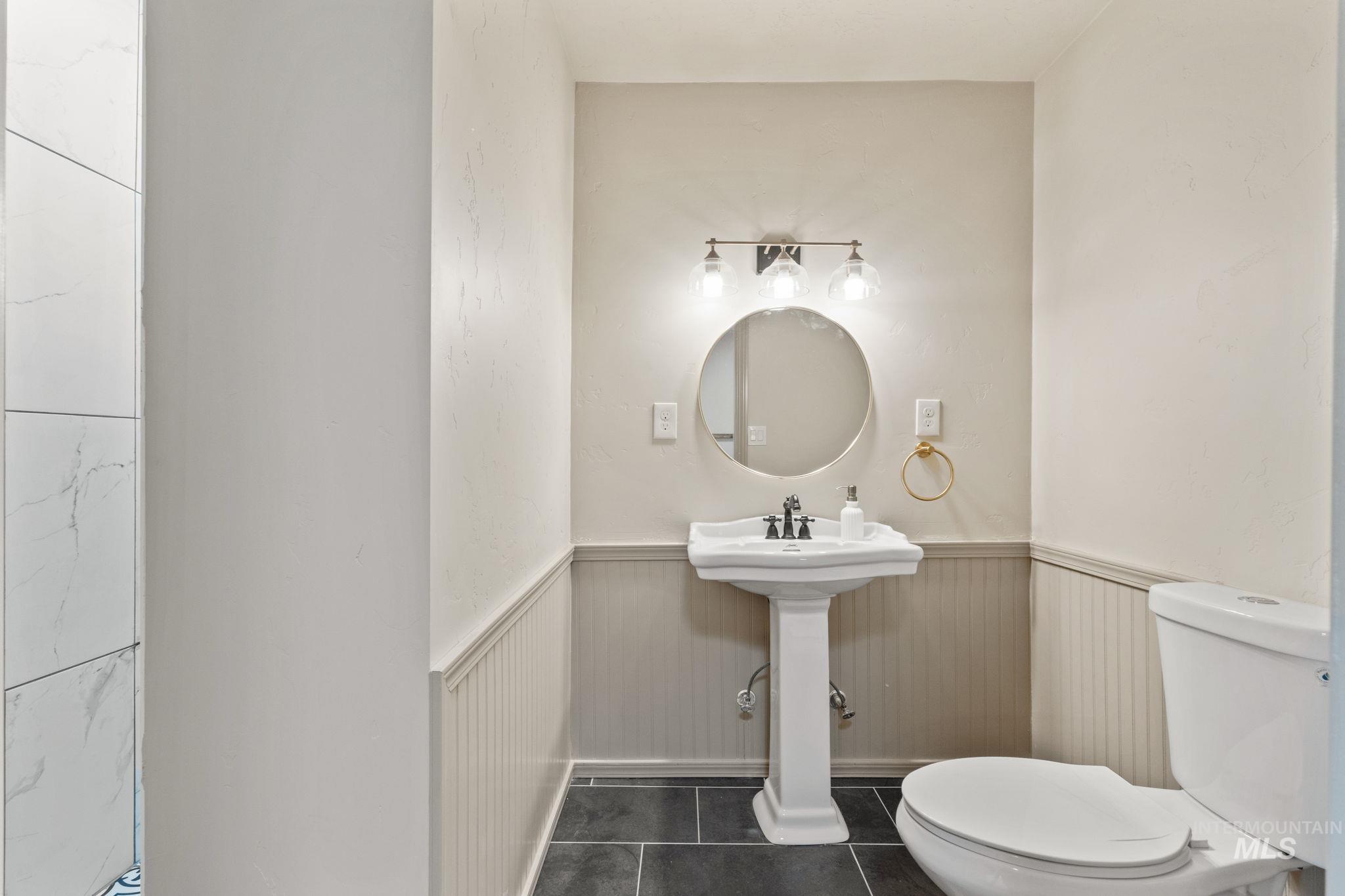 Pedestal sink with a circular mirror, three-light vanity fixture, beadboard wainscoting, and dark tile flooring