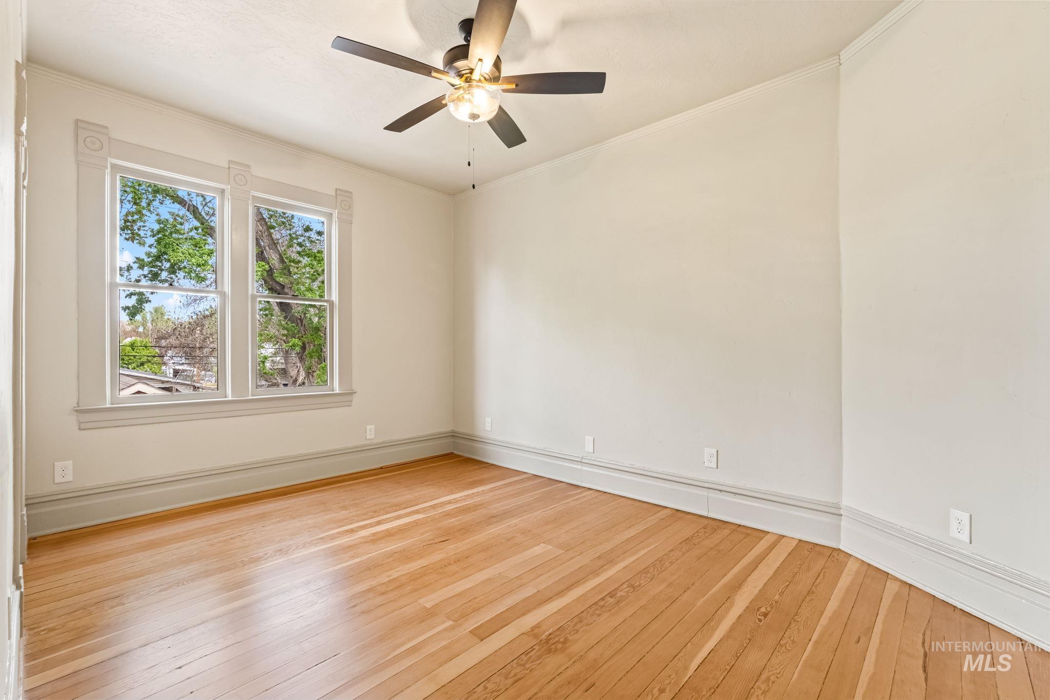 Bright interior space featuring wood-finish flooring, a ceiling fan with integrated lighting, and a large window with decorative trim