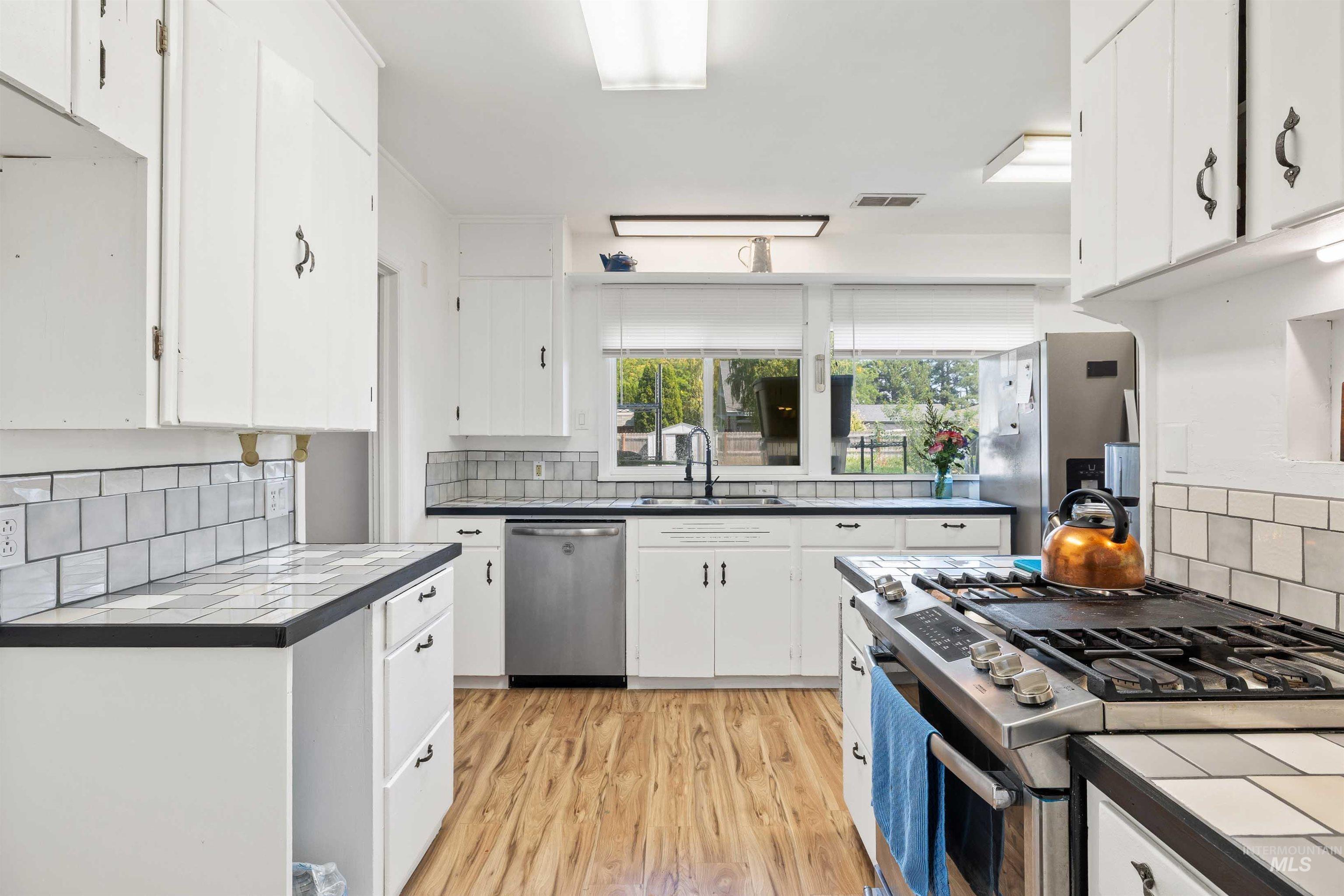 Kitchen with tile counters, stainless steel appliances, white cabinets, backsplash, and light wood finished floors