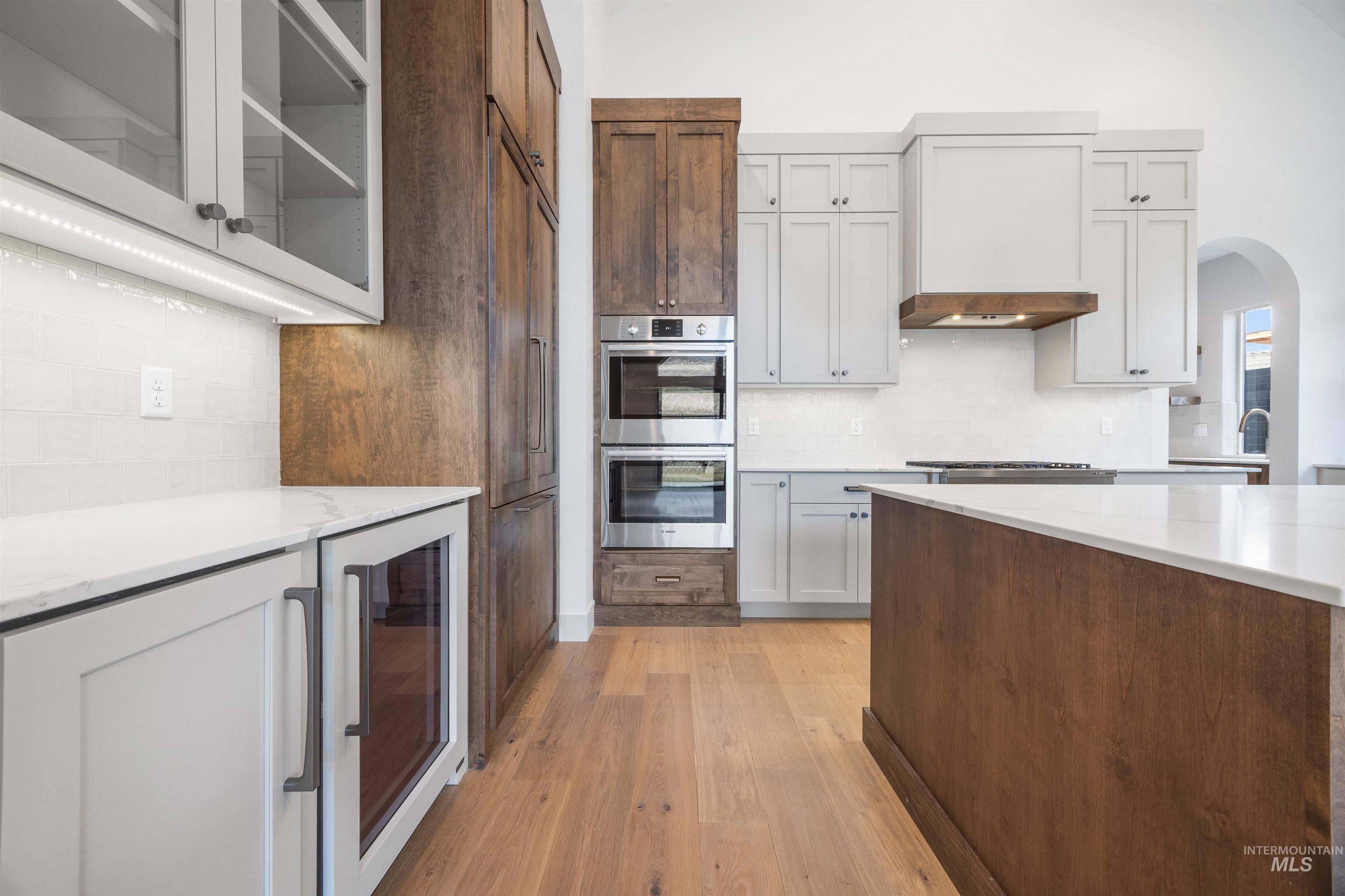 Two tone kitchen with wine cooler, backsplash, arched walkways, light wood-style floors, and stainless steel double oven