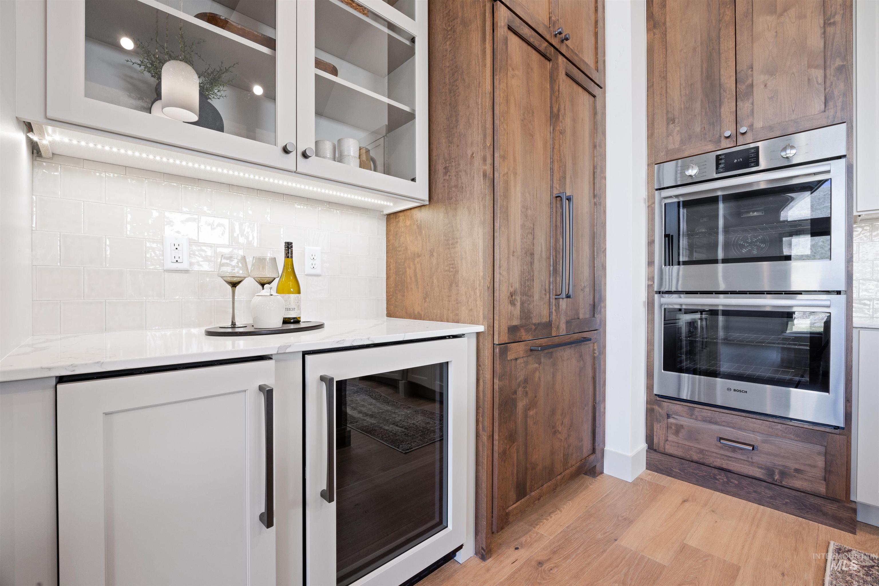 Kitchen with double oven, beverage cooler, decorative backsplash, light stone countertops, and glass fronted cabinets