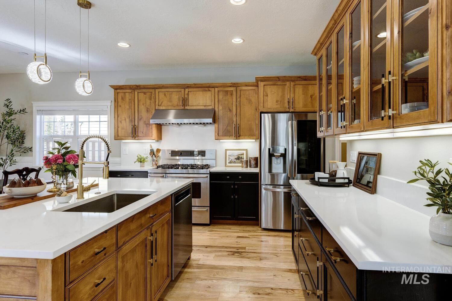 Kitchen with brown cabinetry, decorative light fixtures, appliances with stainless steel finishes, glass insert cabinets, and an island with sink