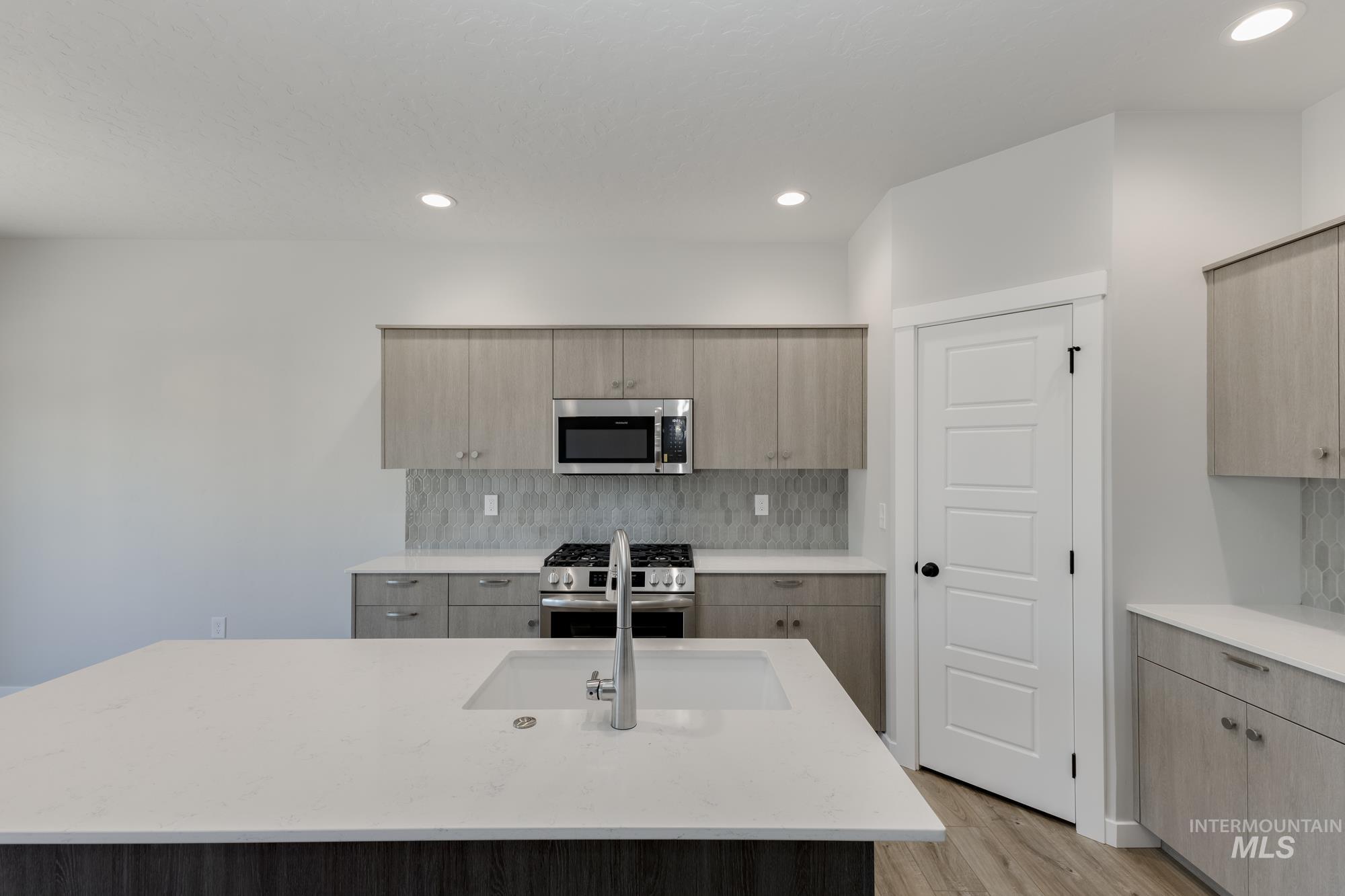 Kitchen with appliances with stainless steel finishes, tasteful backsplash, light wood-type flooring, light stone countertops, and a kitchen island with sink