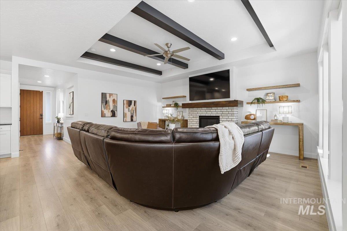Living room featuring a raised ceiling, a fireplace, light wood-style floors, a ceiling fan, and beamed ceiling