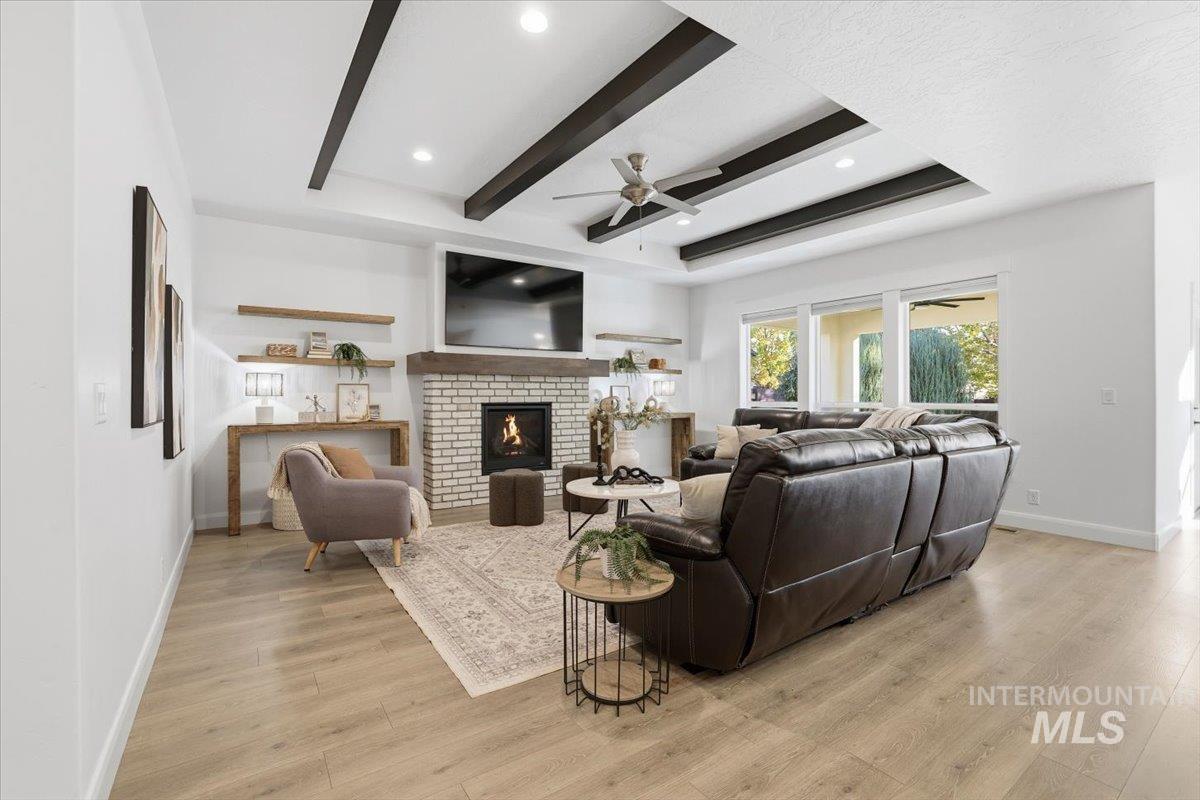 Living area featuring beamed ceiling, a fireplace, light wood-style floors, a ceiling fan, and a tray ceiling