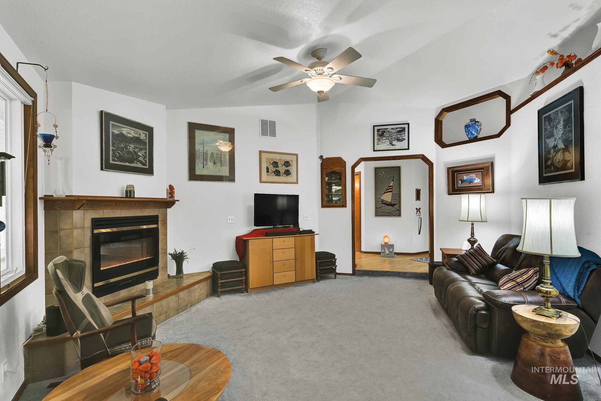 Carpeted living room with a tiled fireplace, vaulted ceiling, a ceiling fan, and arched walkways
