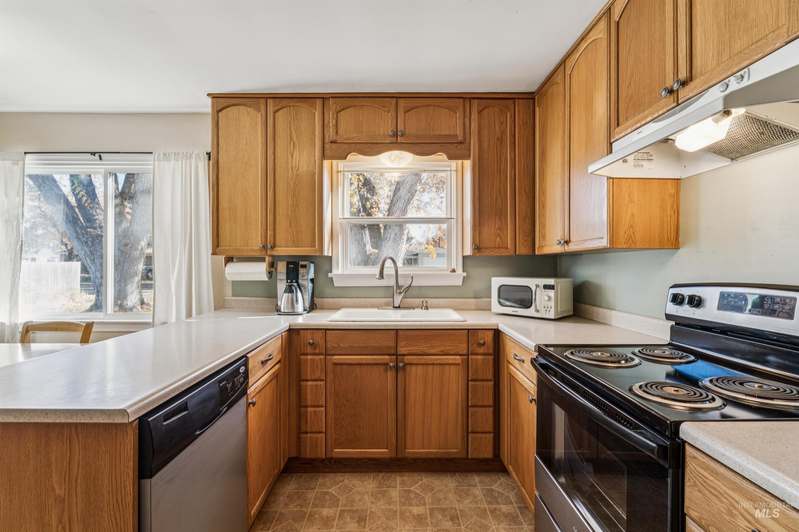 Kitchen with black range with electric stovetop, brown cabinetry, stainless steel dishwasher, under cabinet range hood, and white microwave