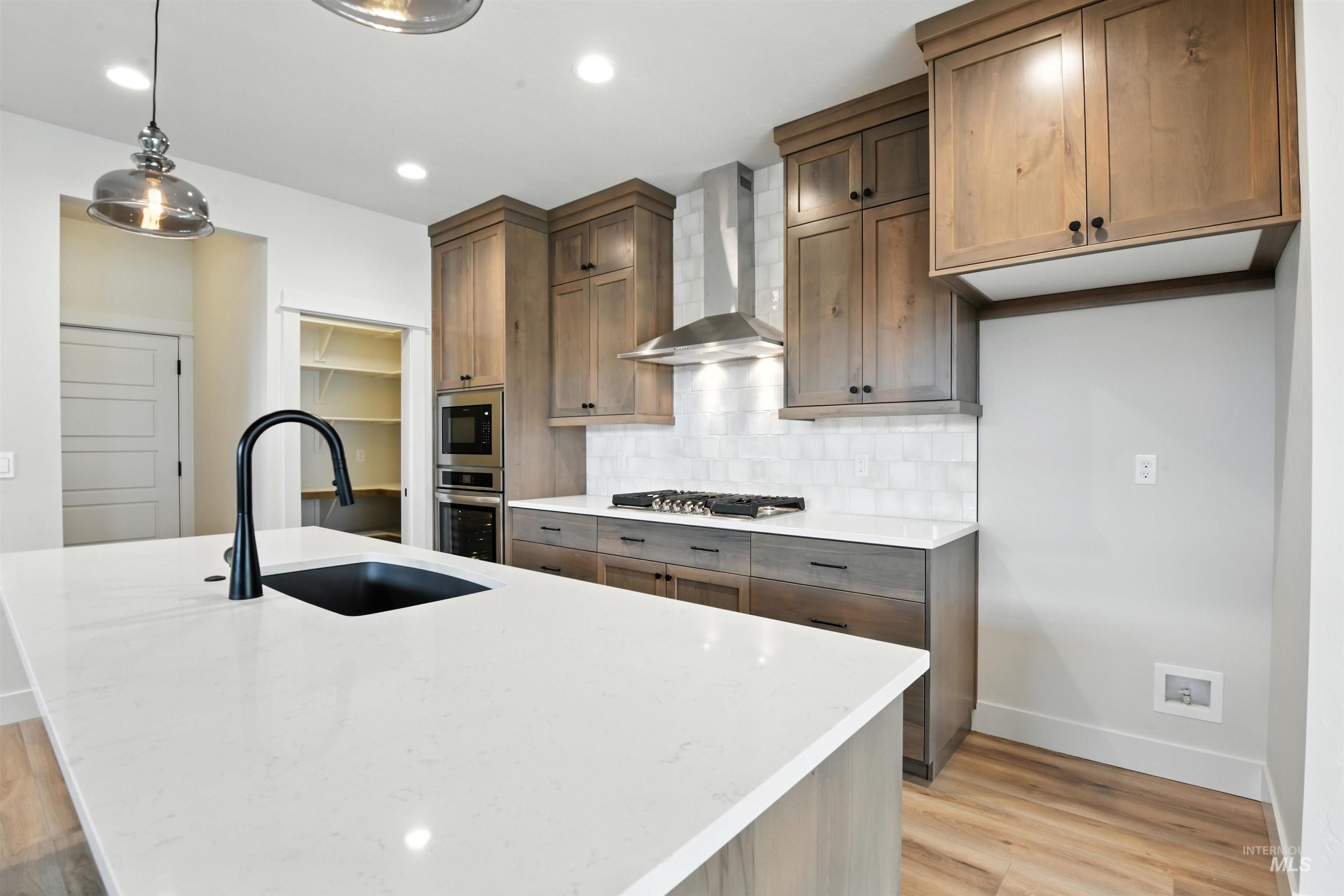 Kitchen featuring wall chimney range hood, stainless steel appliances, light wood finished floors, tasteful backsplash, and a center island with sink