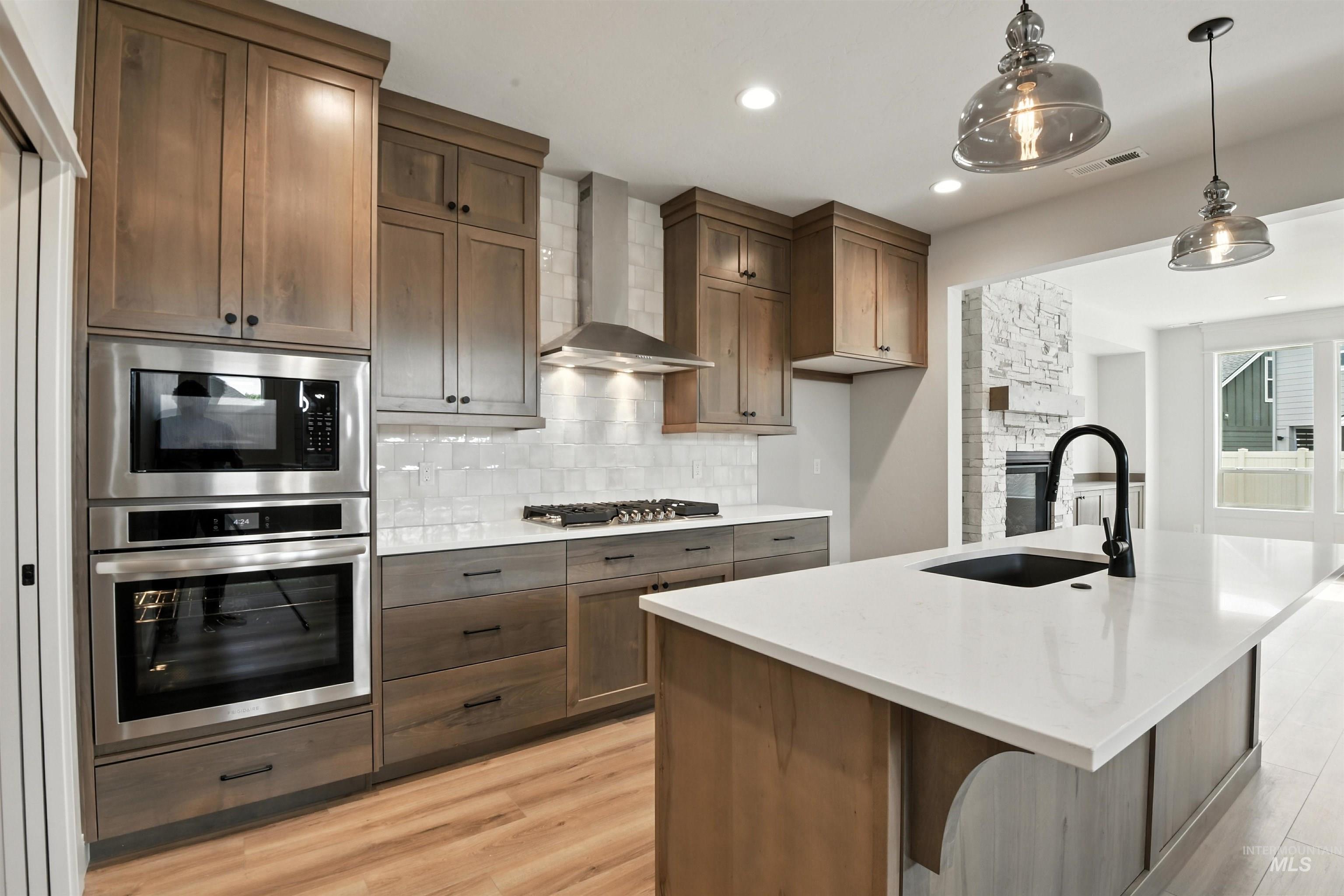 Kitchen with appliances with stainless steel finishes, wall chimney range hood, backsplash, light wood-type flooring, and pendant lighting