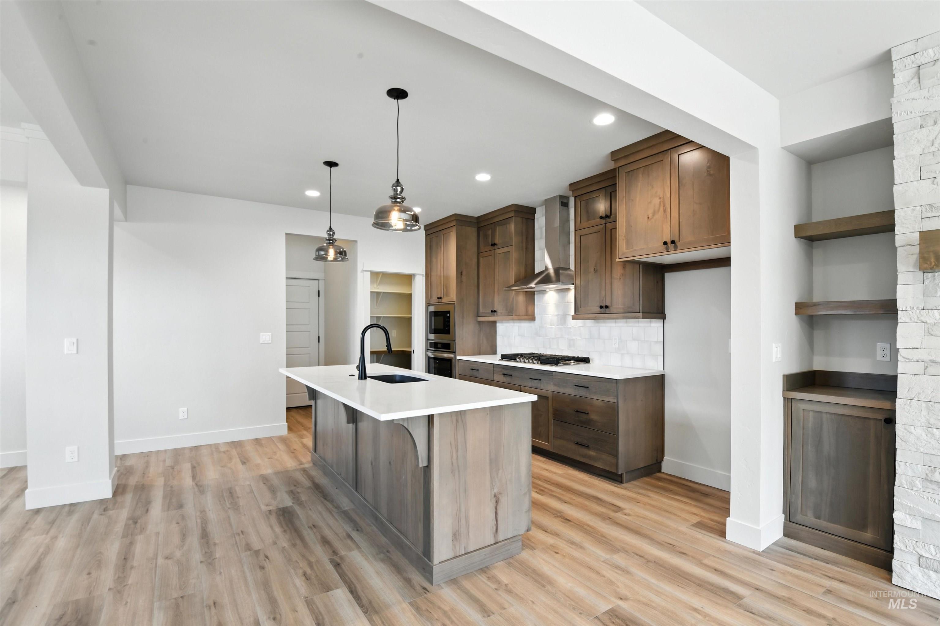 Kitchen with wall chimney exhaust hood, light countertops, decorative backsplash, an island with sink, and open shelves