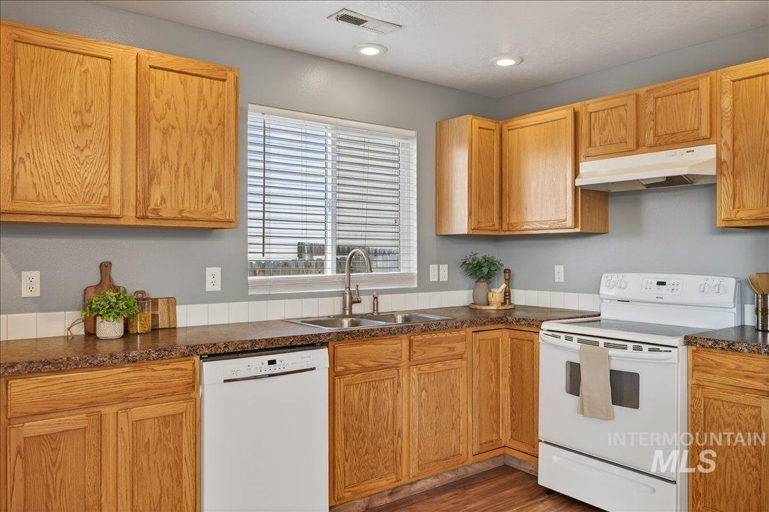 Kitchen with white appliances, under cabinet range hood, dark countertops, and wood finished floors