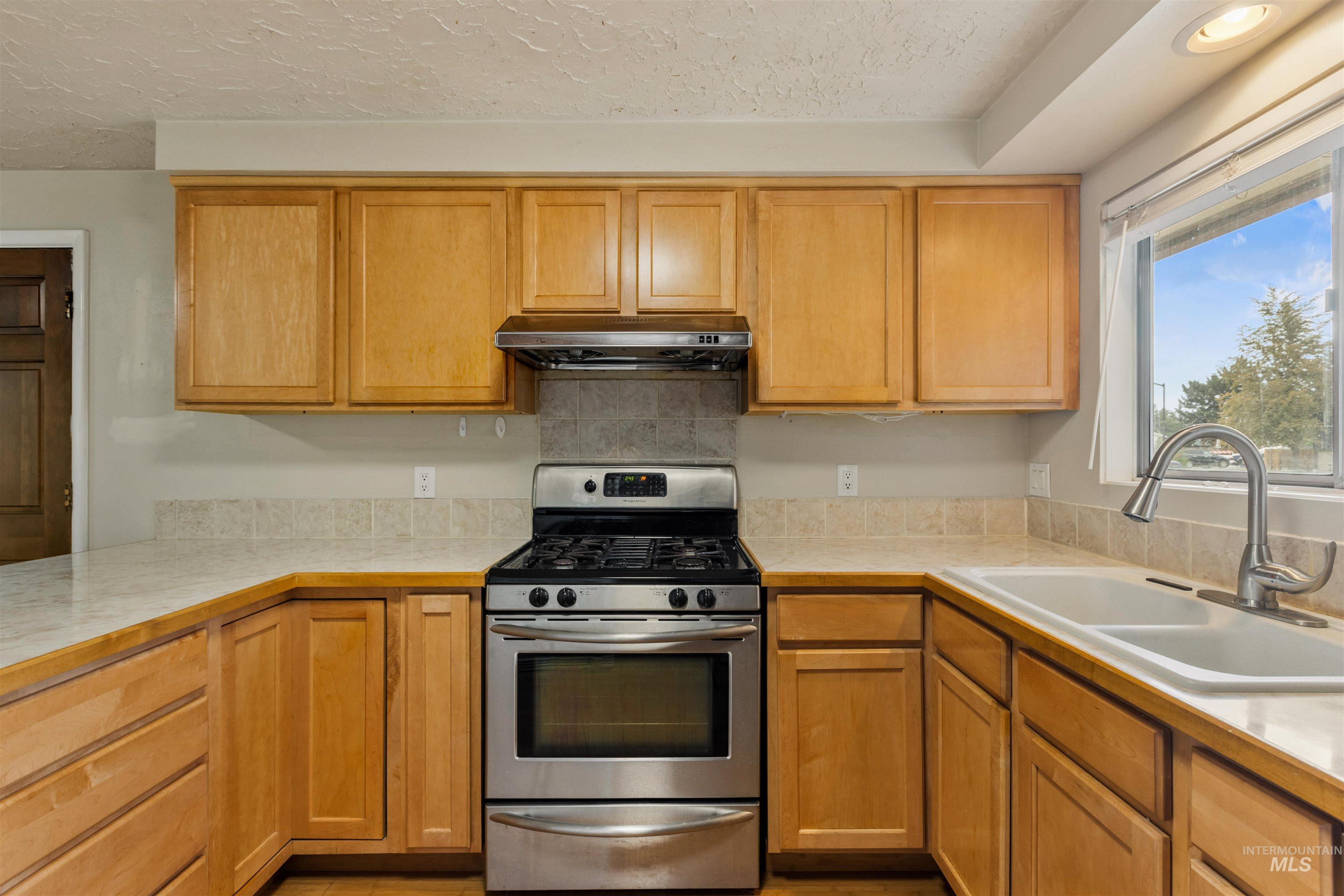 Kitchen with stainless steel gas range oven, light countertops, under cabinet range hood, a textured ceiling, and brown cabinets