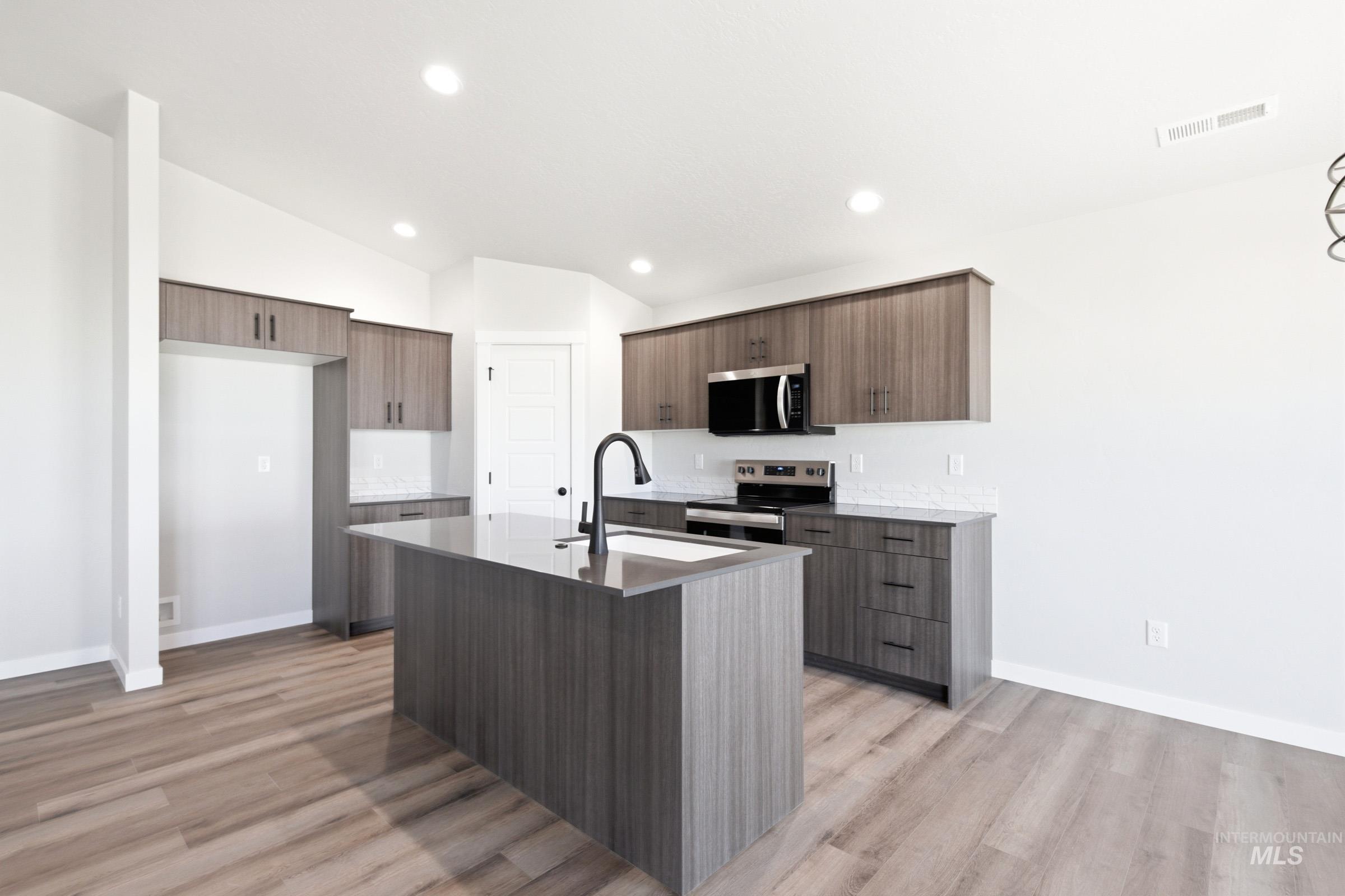 Open-concept kitchen featuring an oversized island with a built-in sink and matte black faucet, wood-finish flooring, recessed lighting, white subway tile backsplash, and stainless steel appliances