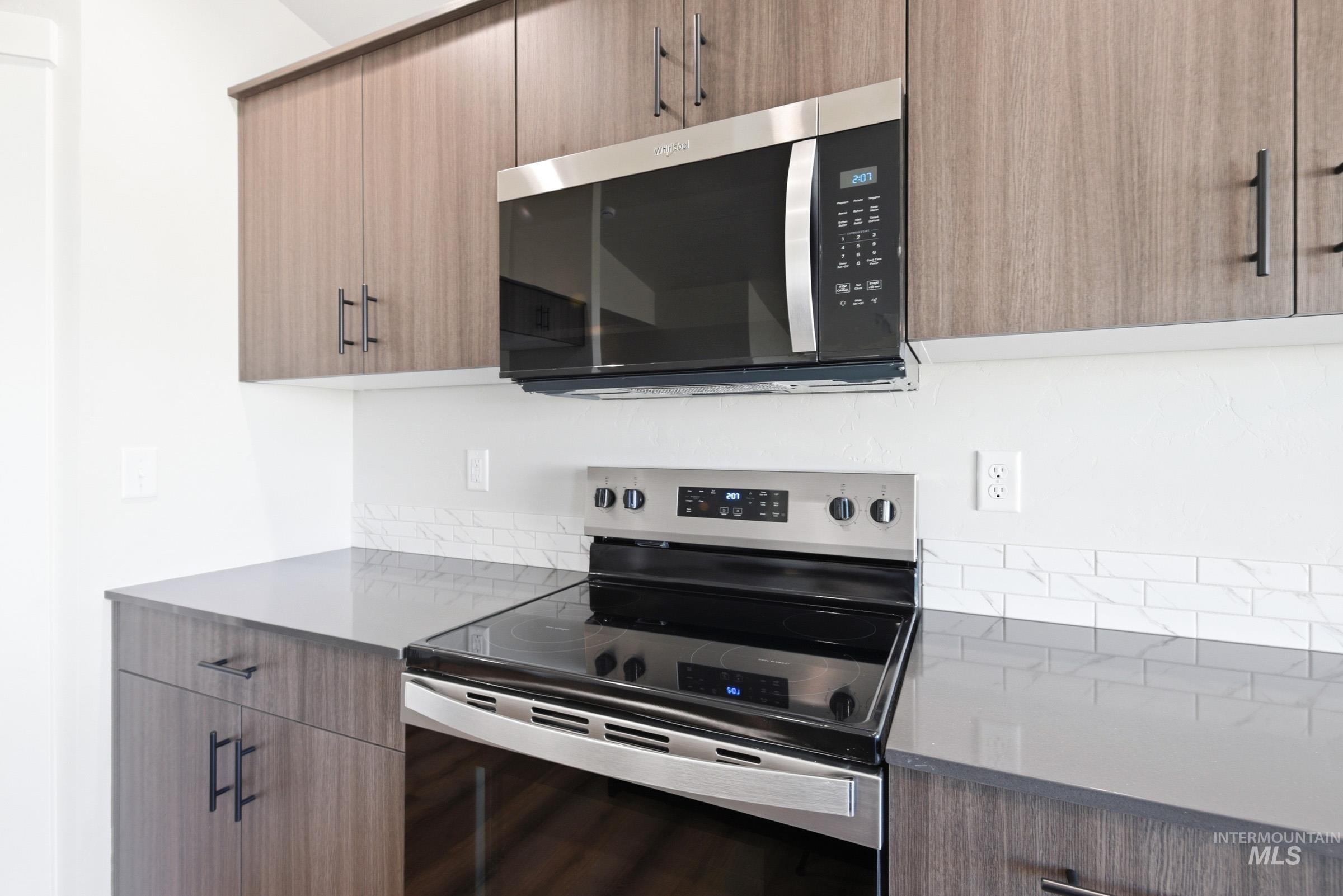Modern kitchen featuring stainless steel appliances, dark wood-finish cabinetry, sleek dark countertops, and a white subway tile backsplash