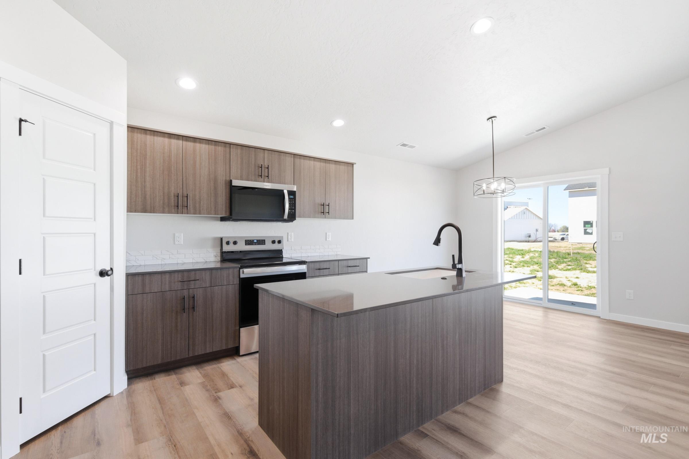 Open-concept kitchen featuring wood-finish cabinetry, stainless steel appliances, a center island with a black faucet, and a tile backsplash