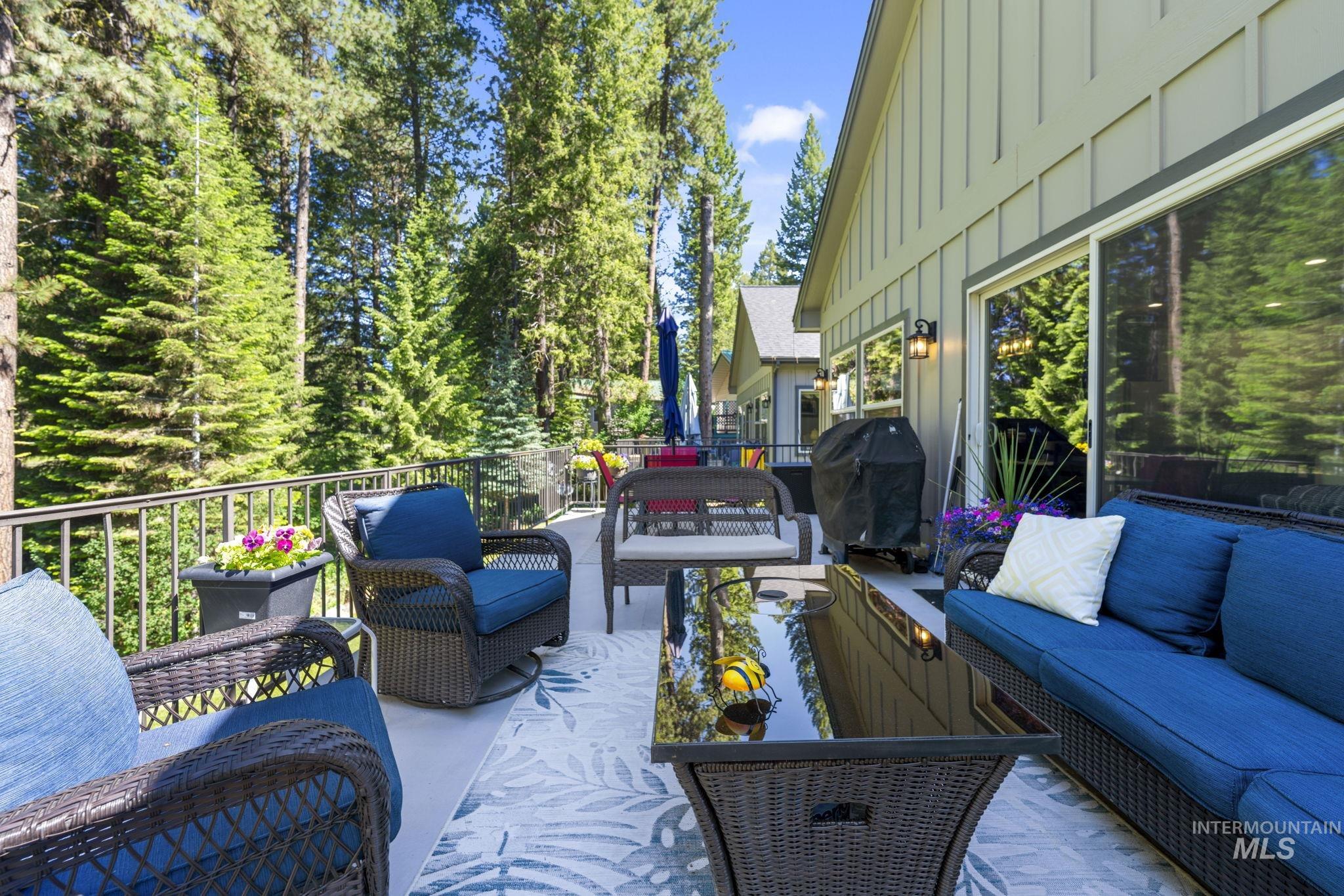 Spacious outdoor deck featuring a patterned area rug, a dark metal railing, and a backdrop of mature evergreen trees