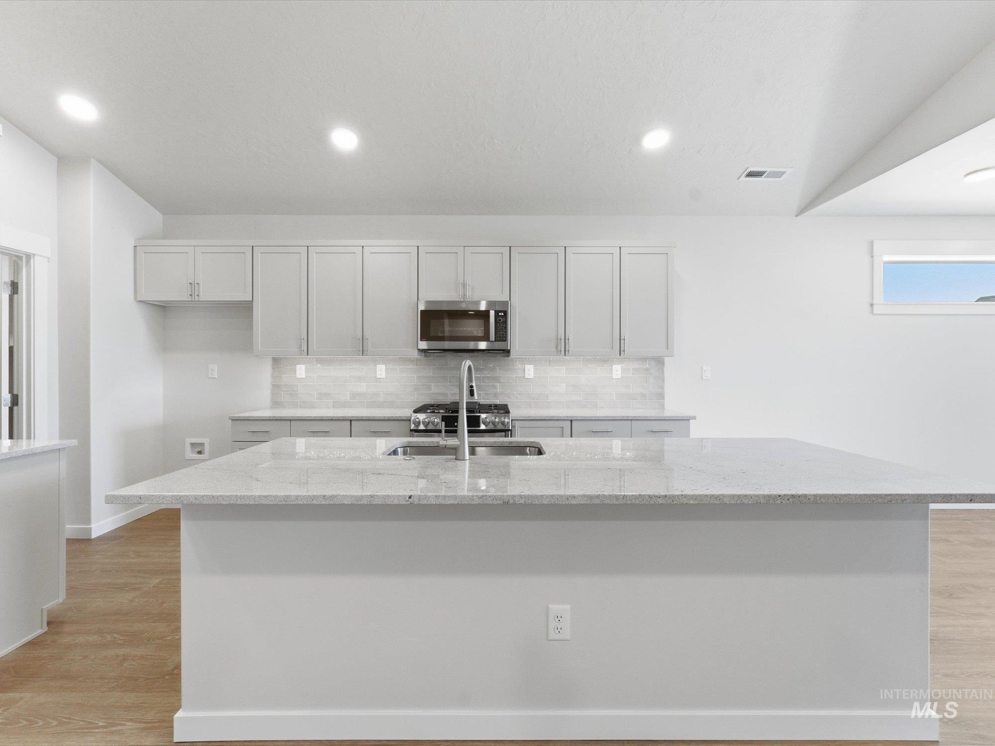 Kitchen featuring an expansive island with a light-colored countertop, recessed lighting, wood-finish flooring, light gray cabinetry, and stainless steel appliances including an oven range, microwave, and sink with a pull-down faucet