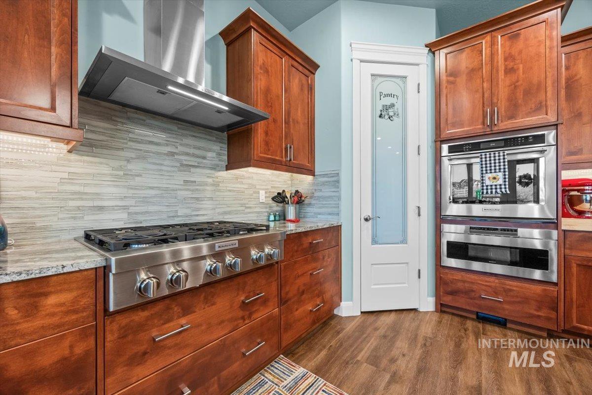 Kitchen with wall chimney range hood, tasteful backsplash, stainless steel appliances, dark wood-style floors, and light stone counters