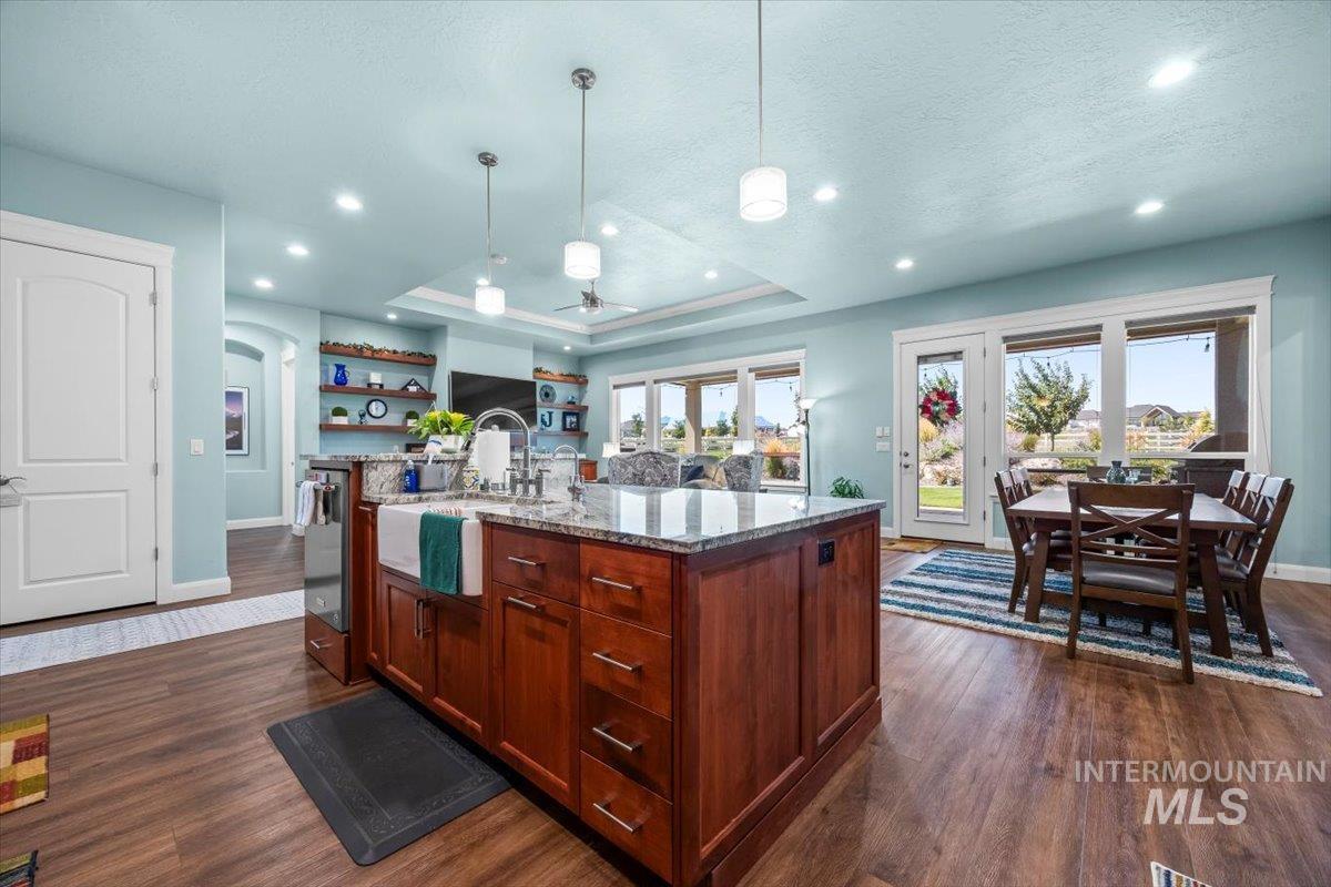 Kitchen with light stone countertops, hanging light fixtures, a raised ceiling, a kitchen island with sink, and dark wood finished floors
