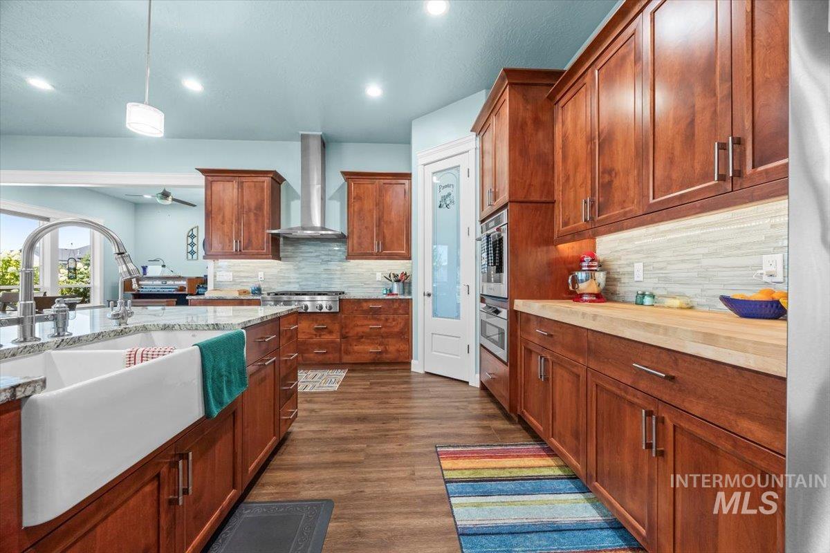 Kitchen with brown cabinetry, dark wood-type flooring, wall chimney exhaust hood, hanging light fixtures, and a ceiling fan