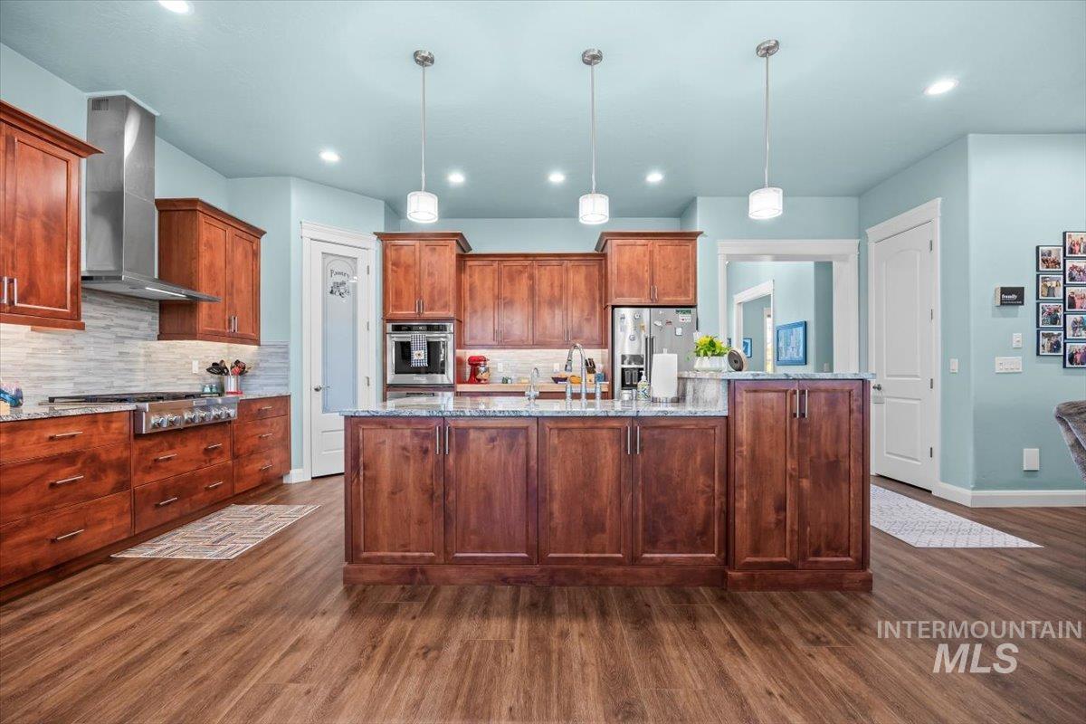 Kitchen featuring a center island with sink, light stone counters, hanging light fixtures, wall chimney range hood, and recessed lighting