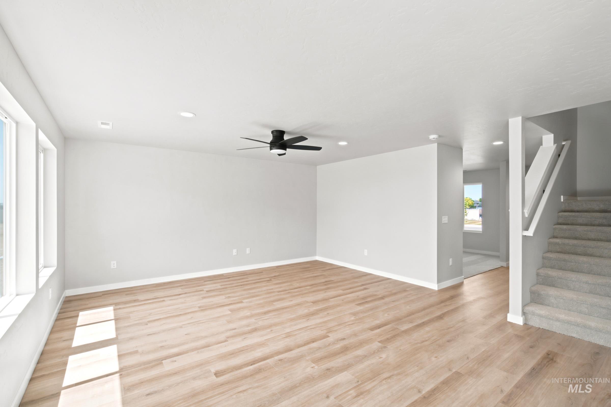 Spacious living area featuring light wood-finish flooring, recessed lighting, a ceiling fan with black blades, multiple windows, and a carpeted staircase with a contemporary white railing