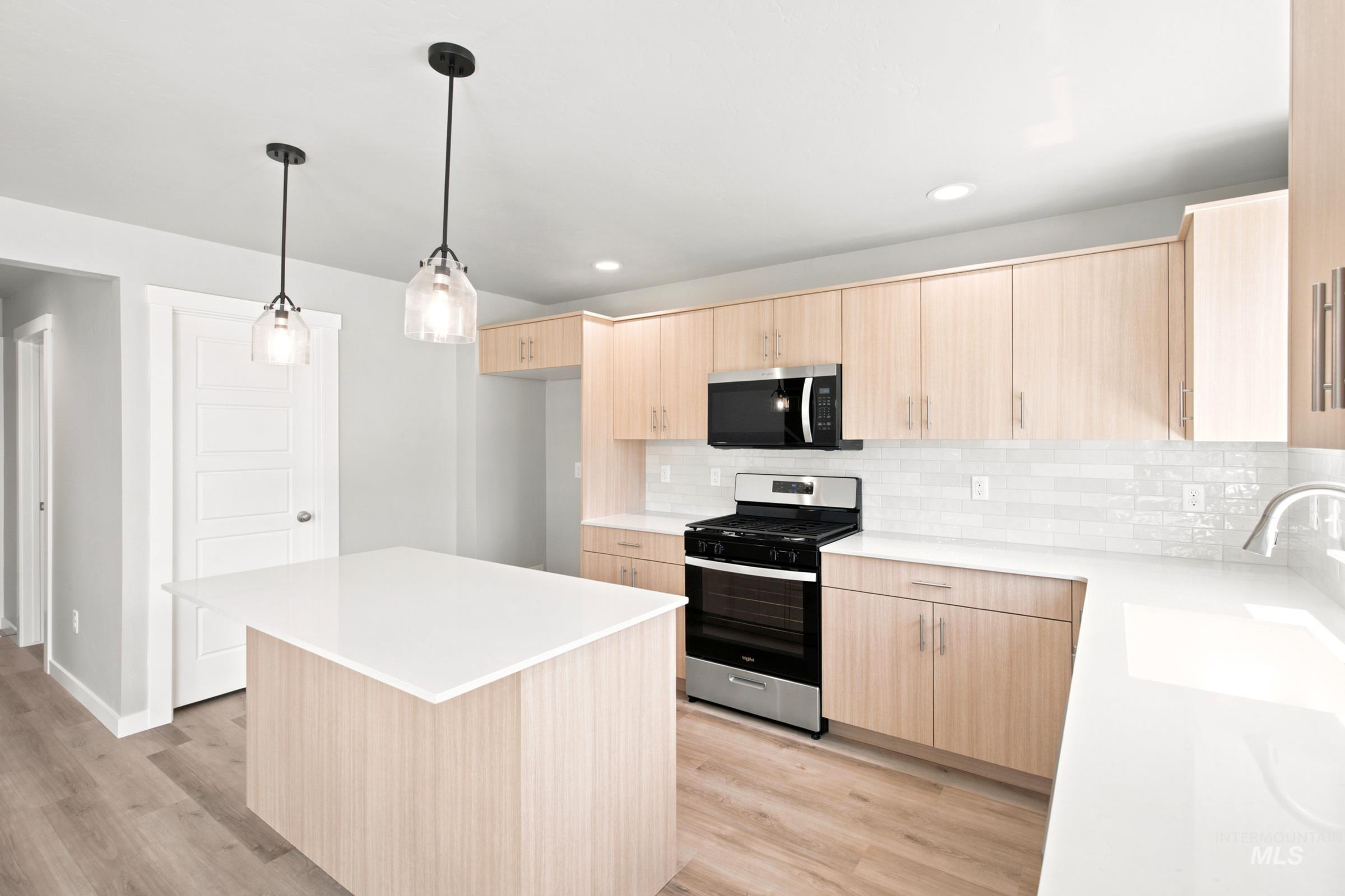 Modern kitchen featuring a central island with white countertop, pendant lighting, light wood-finish cabinetry, subway tile backsplash, and stainless steel appliances