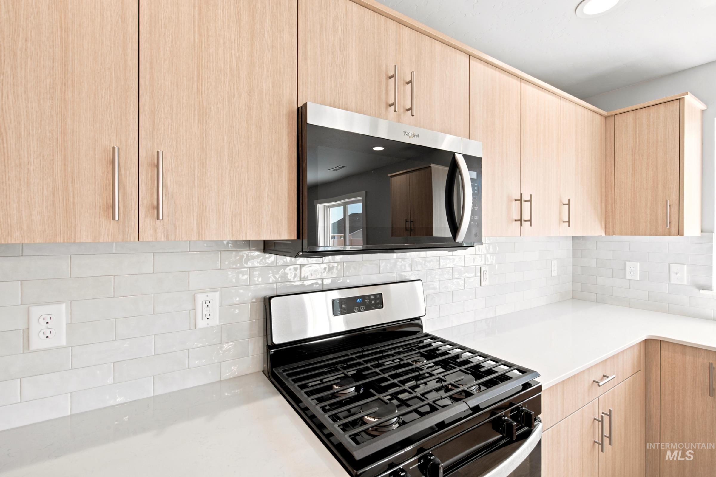 Kitchen featuring light wood-finish cabinetry, stainless steel appliances including a microwave and gas range, white subway tile backsplash, and light-toned countertops