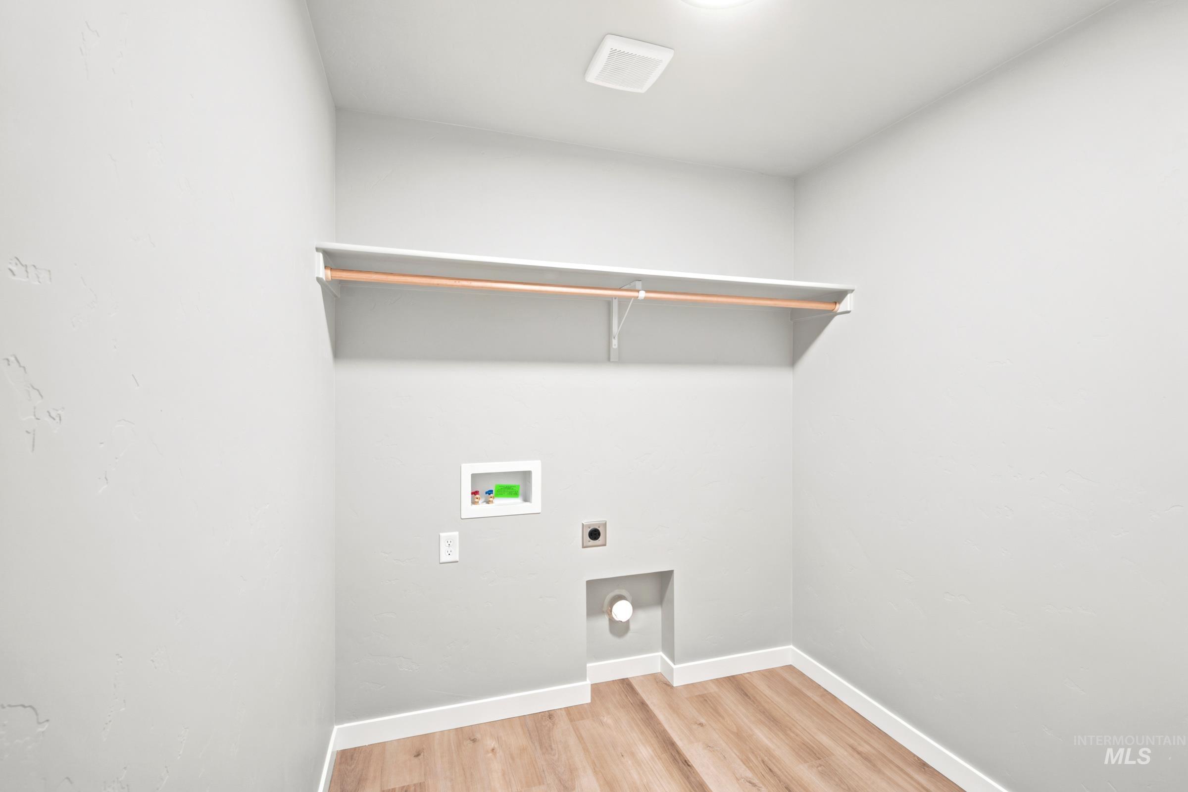 Dedicated utility space featuring light wood-finish flooring, a white baseboard, a ceiling-mounted light fixture, and a wall-mounted shelving unit with hanging rod