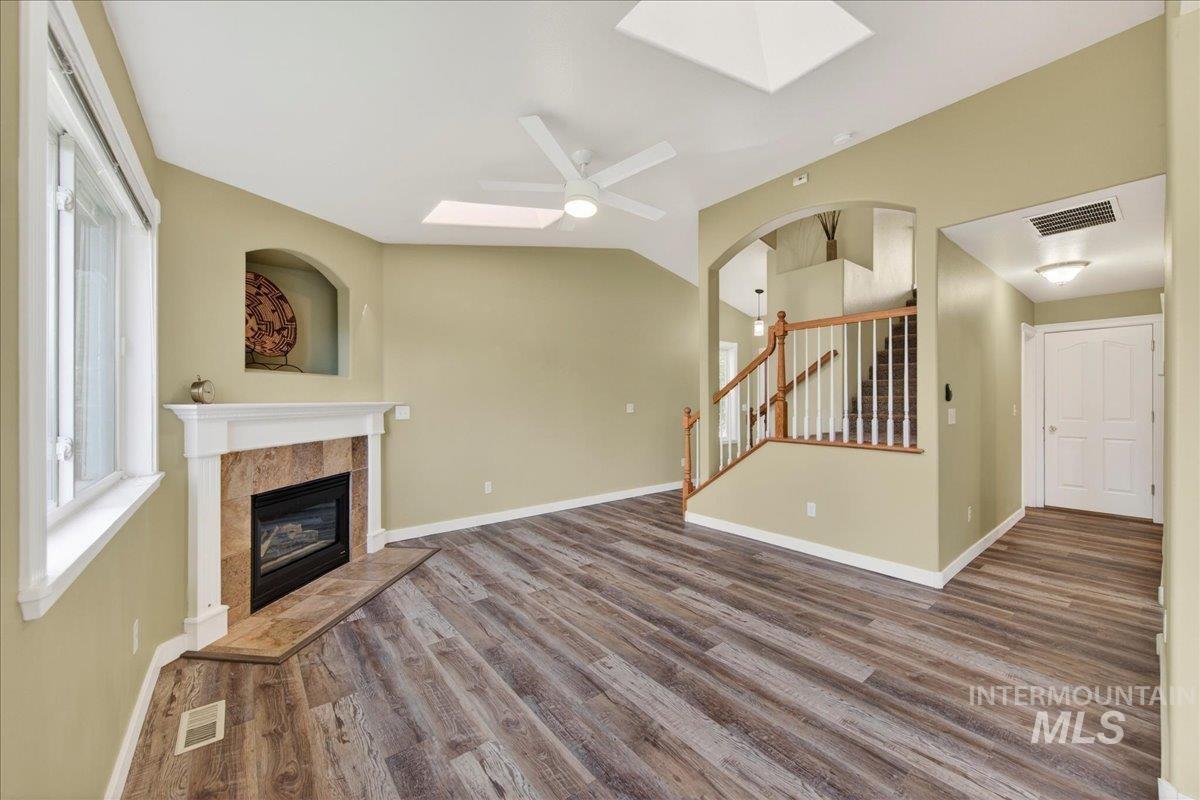 Unfurnished living room featuring a skylight, a tiled fireplace, dark wood-style floors, ceiling fan, and vaulted ceiling