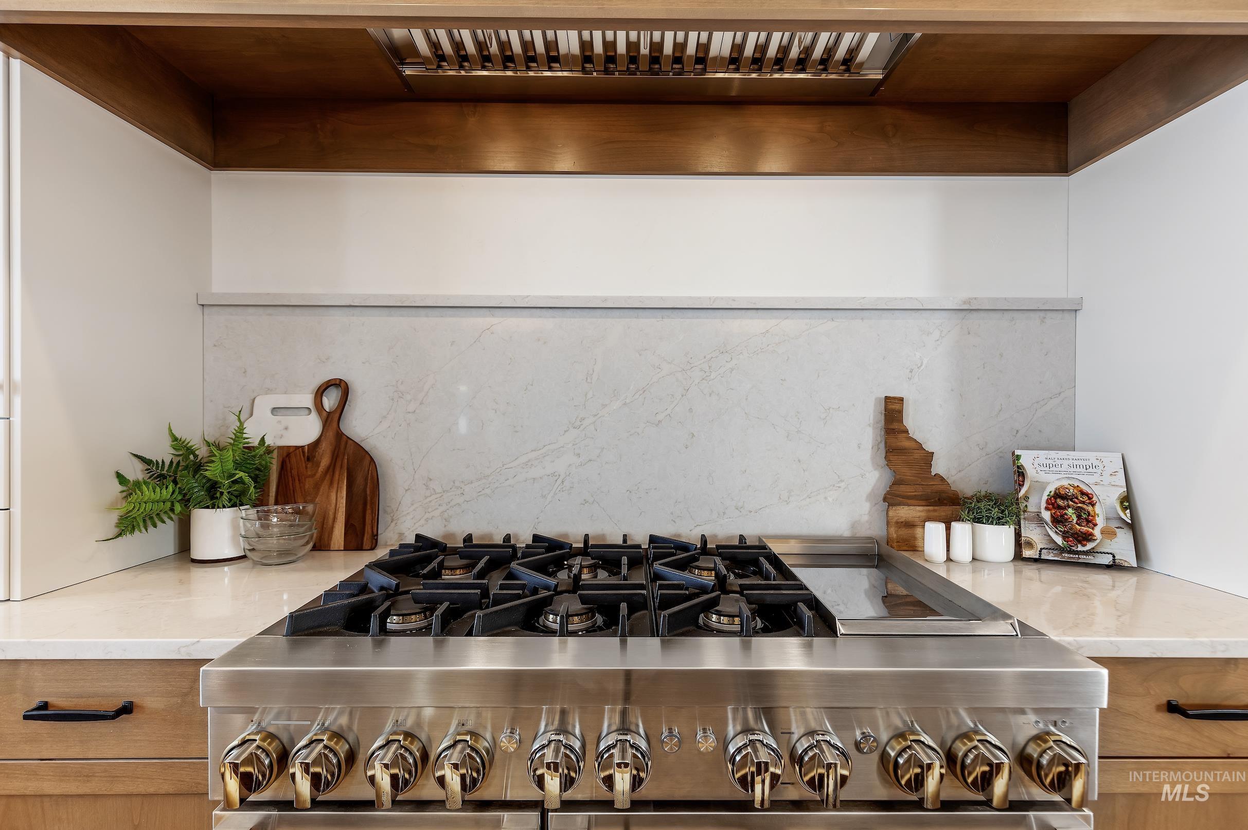 Kitchen view of light stone countertops, range hood, backsplash, brown cabinetry, and high end stove