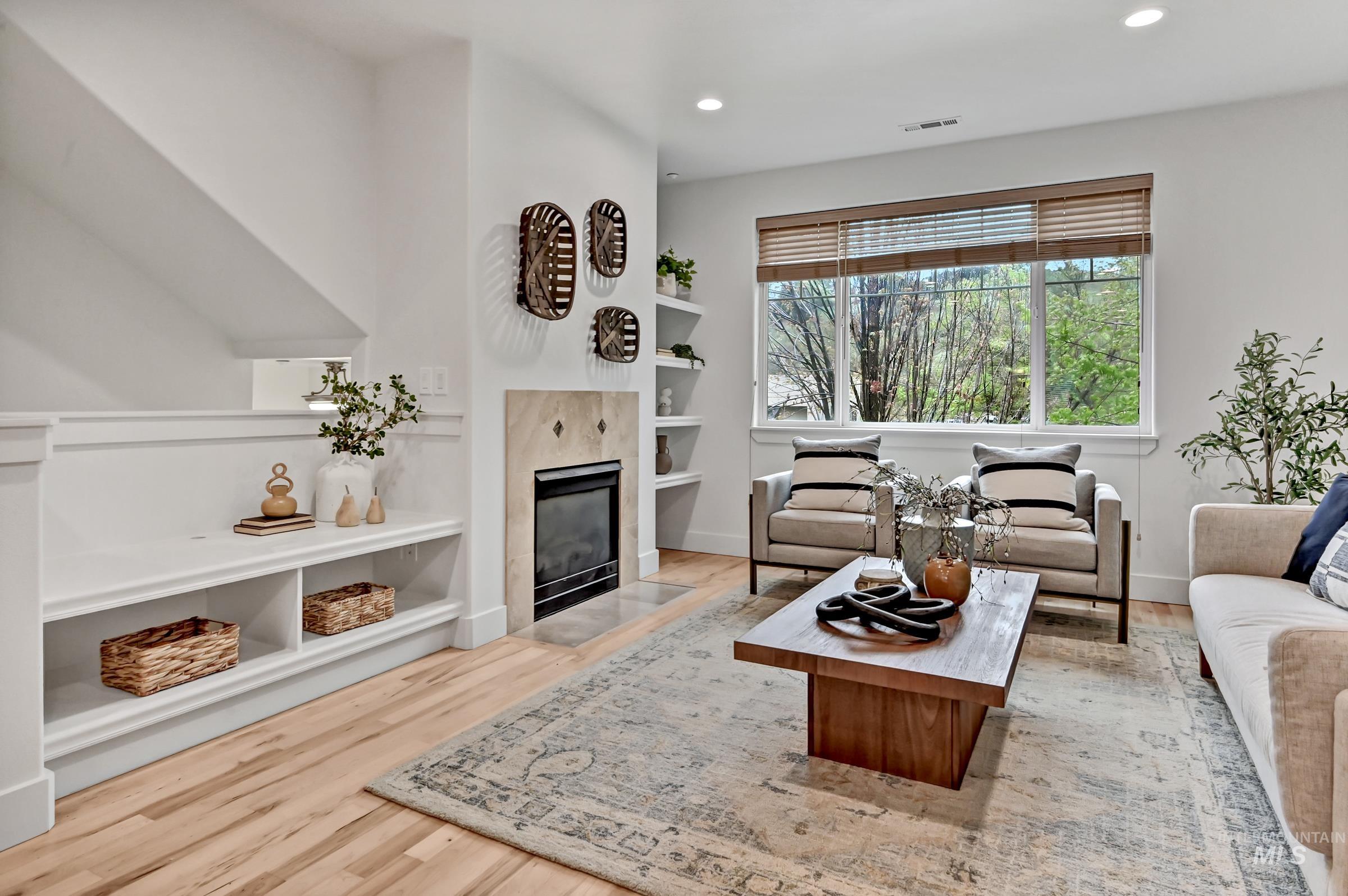 Living room with light walls, hardwood floors, and large front window. Bright space with comfortable seating area and gas fireplace.