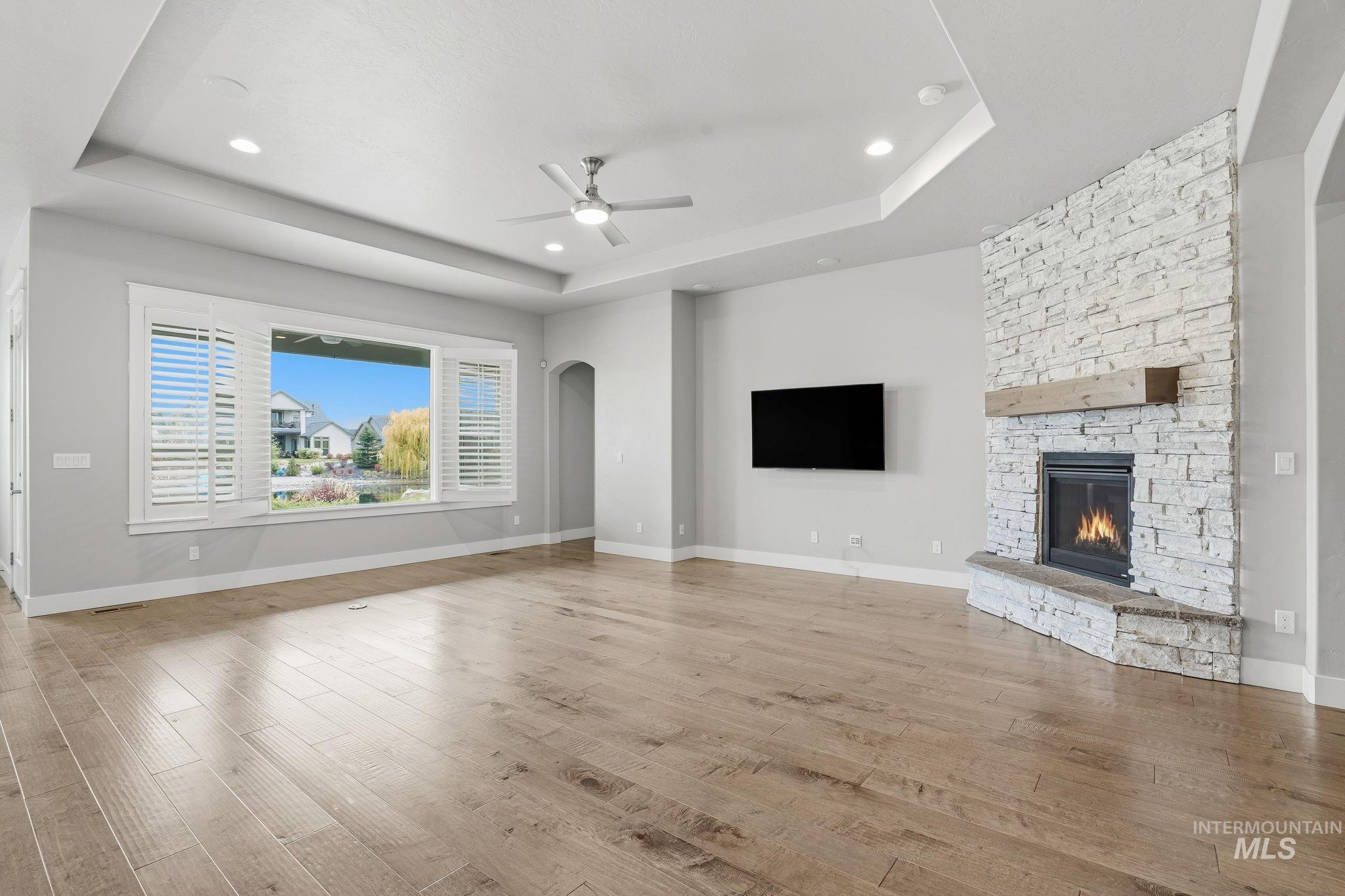 Unfurnished living room with arched walkways, light wood-style floors, a raised ceiling, a stone fireplace, and recessed lighting