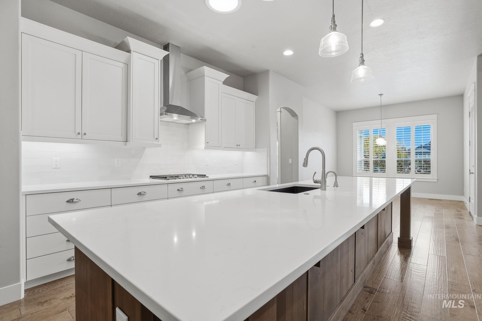 Kitchen with light wood finished floors, tasteful backsplash, decorative light fixtures, a large island with sink, and wall chimney exhaust hood