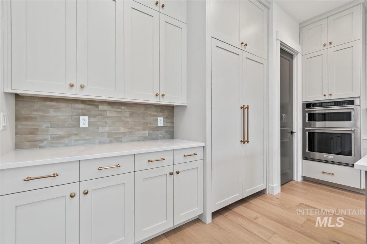 Kitchen featuring white cabinets, light wood-style floors, double oven, decorative backsplash, and light stone countertops