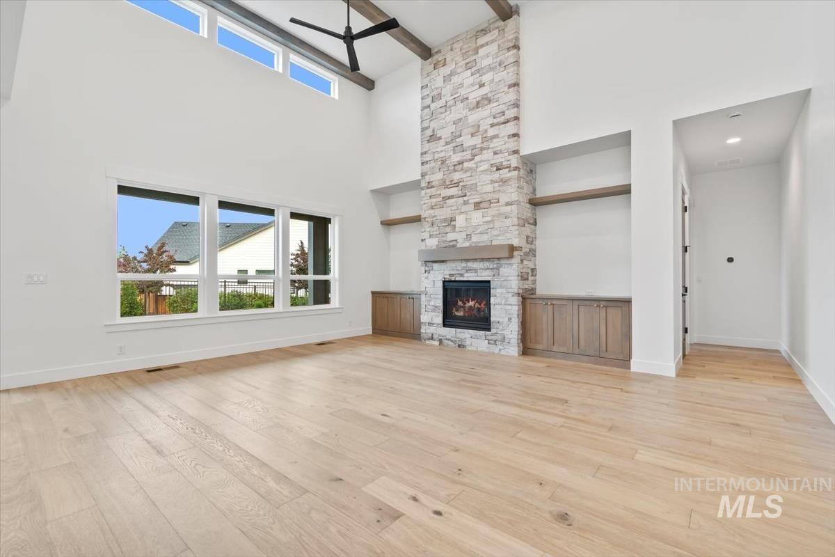 Unfurnished living room with light wood-style floors, a stone fireplace, a towering ceiling, beam ceiling, and a ceiling fan