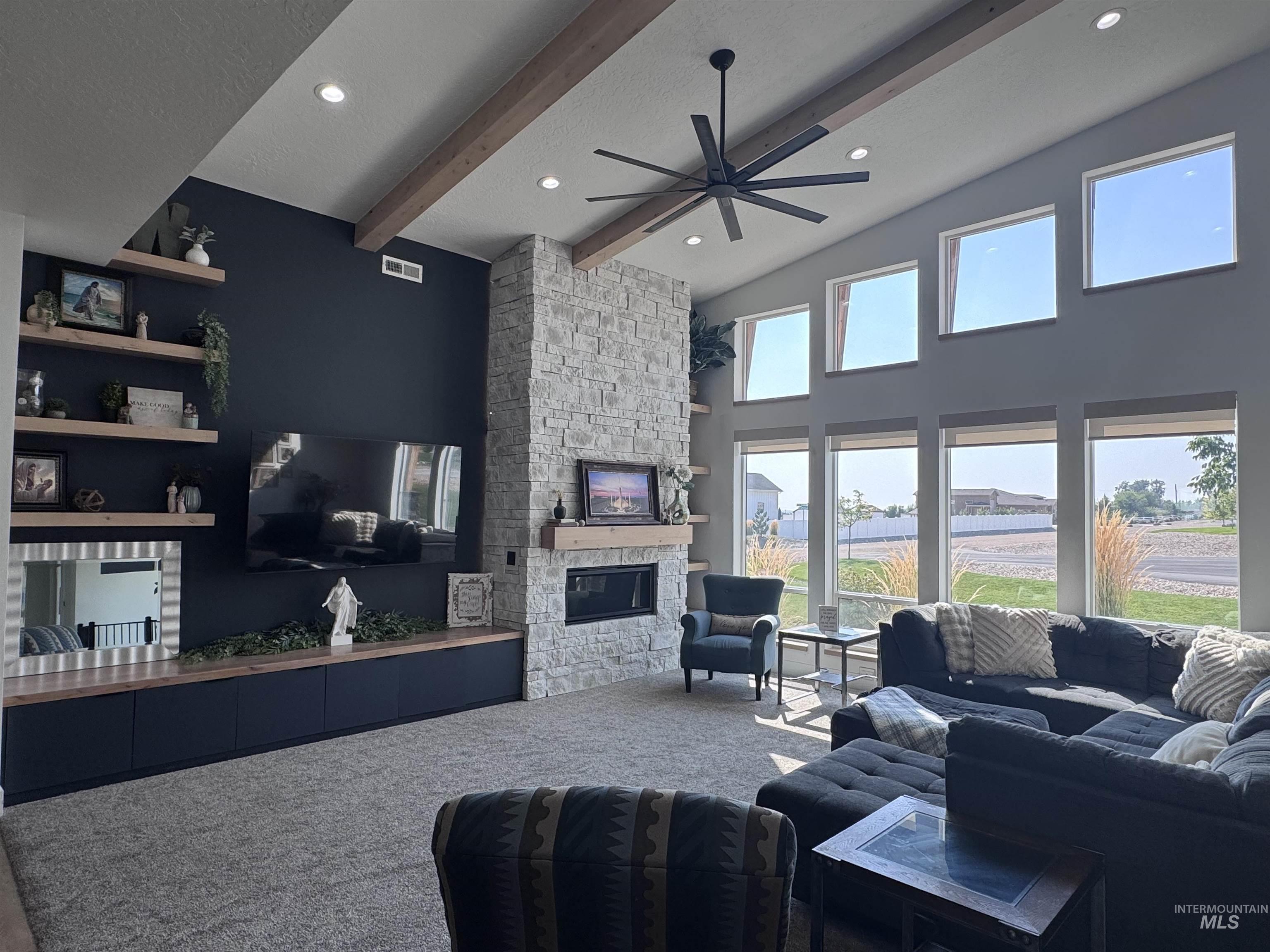 Carpeted living room featuring a stone fireplace, beam ceiling, a ceiling fan, recessed lighting, and a high ceiling