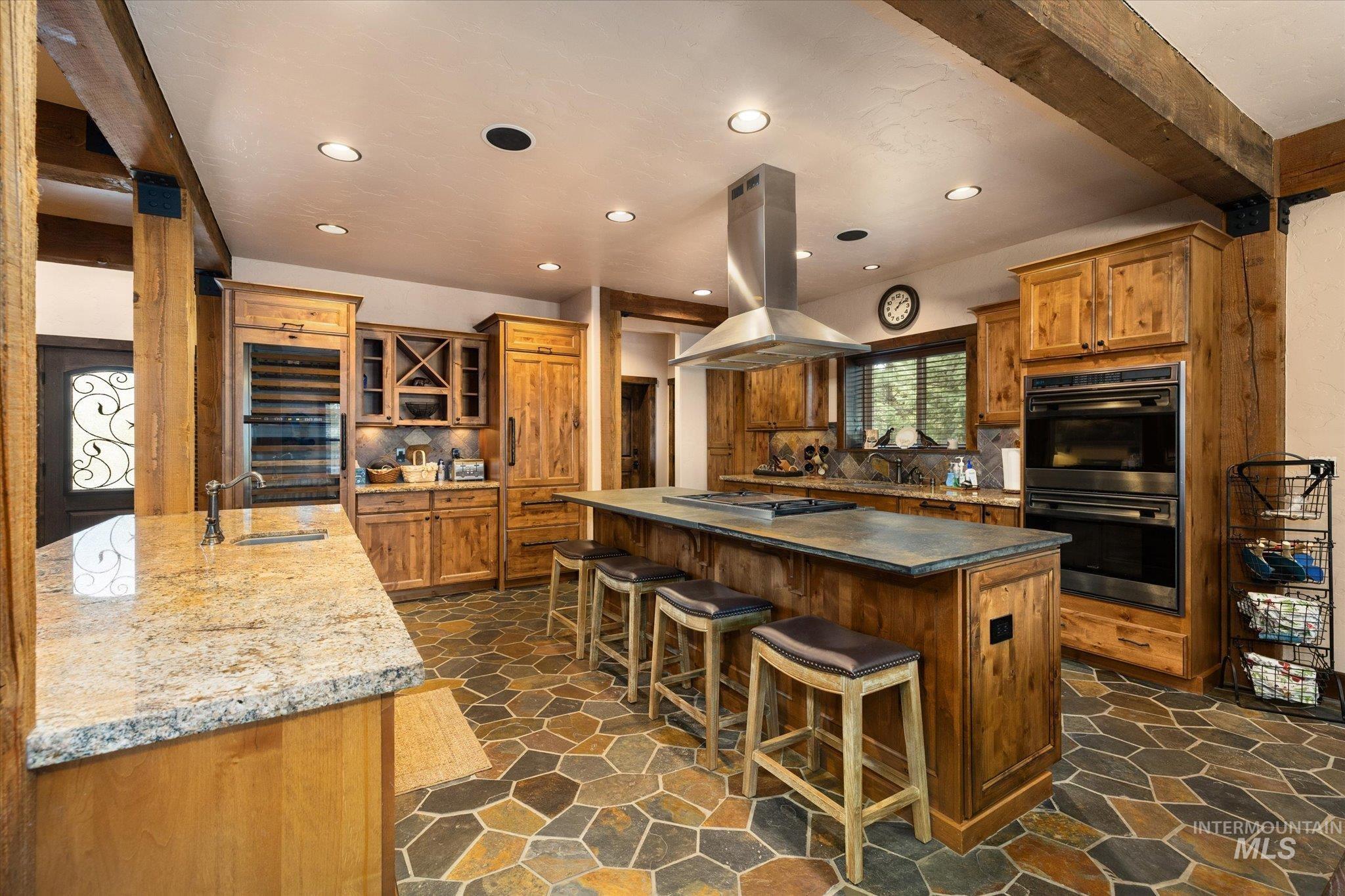 Spacious kitchen featuring natural wood cabinetry, stone tile flooring, a large central island with seating, a stainless steel range hood, and integrated double ovens