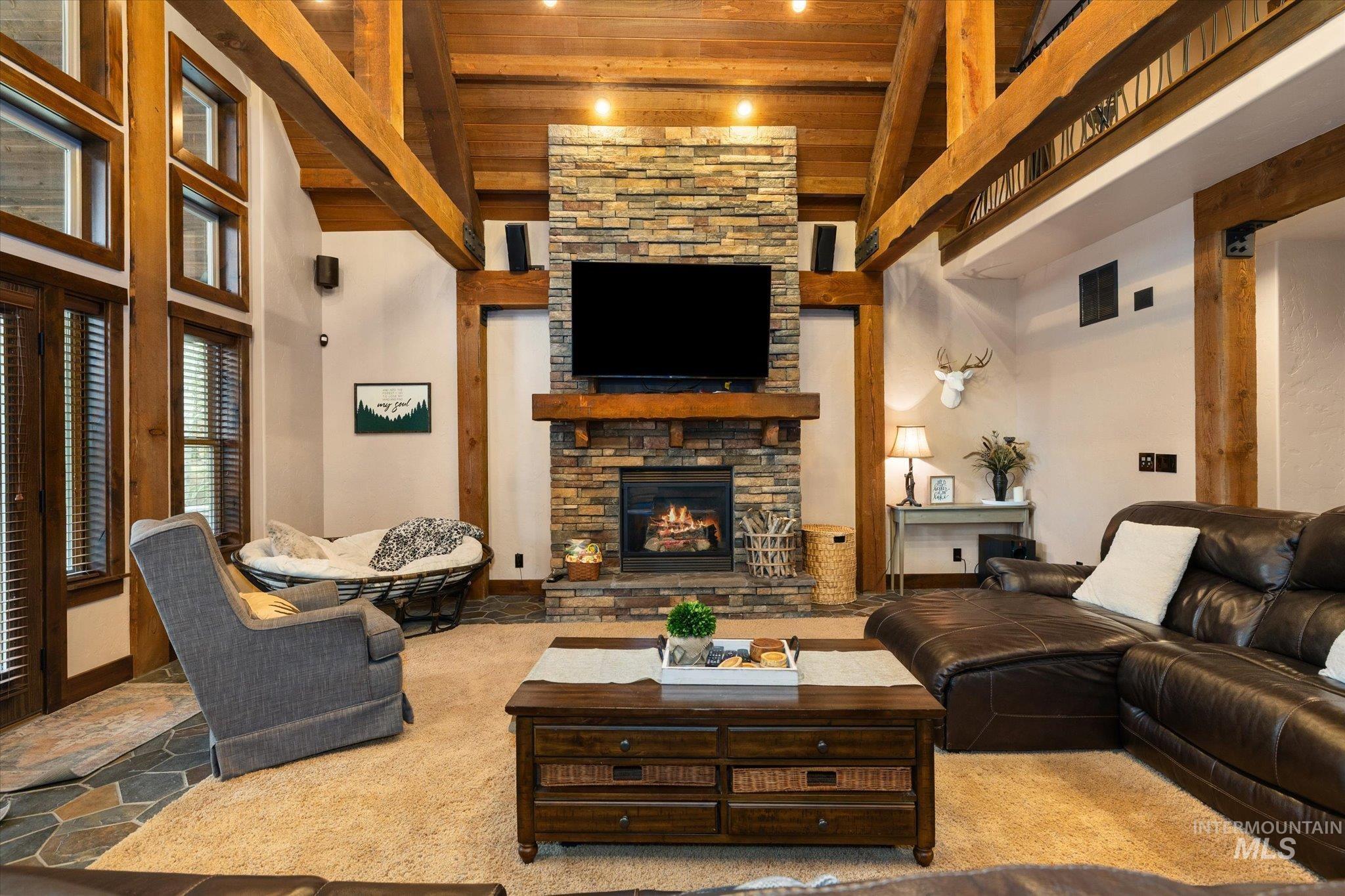 Spacious living area featuring a vaulted wood-beam ceiling, stone-clad fireplace, and large windows with integrated blinds