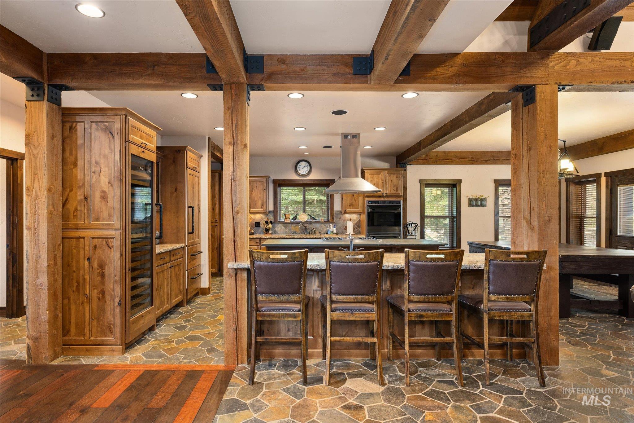 Rustic-inspired kitchen featuring exposed wood beams, stone tile flooring, a large island with seating, integrated wine refrigeration, and stainless steel appliances